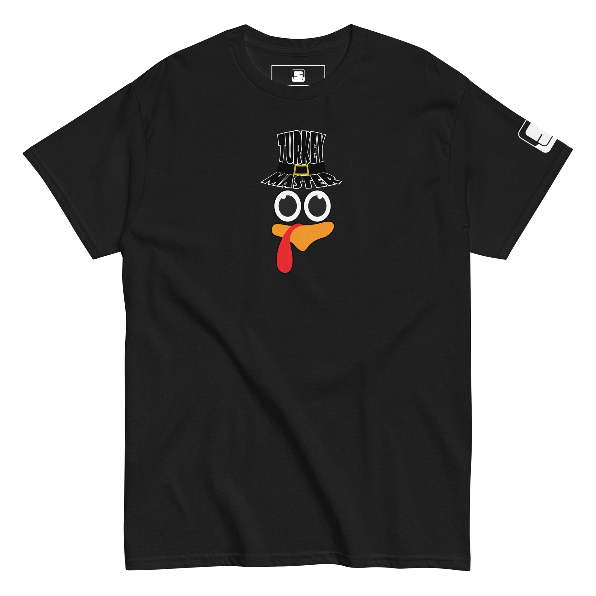 A black t-shirt featuring a playful graphic design of a turkey face with eyes, an orange beak, and a red snood. Above the face, the text reads "TURKEY MASTER" in bold, stylized letters. The text is in the shape of a pilgrims hat. On the left sleeve, there's a rectangular logo patch, adding a unique touch. The shirt is laid out flat, showcasing the entire design clearly.