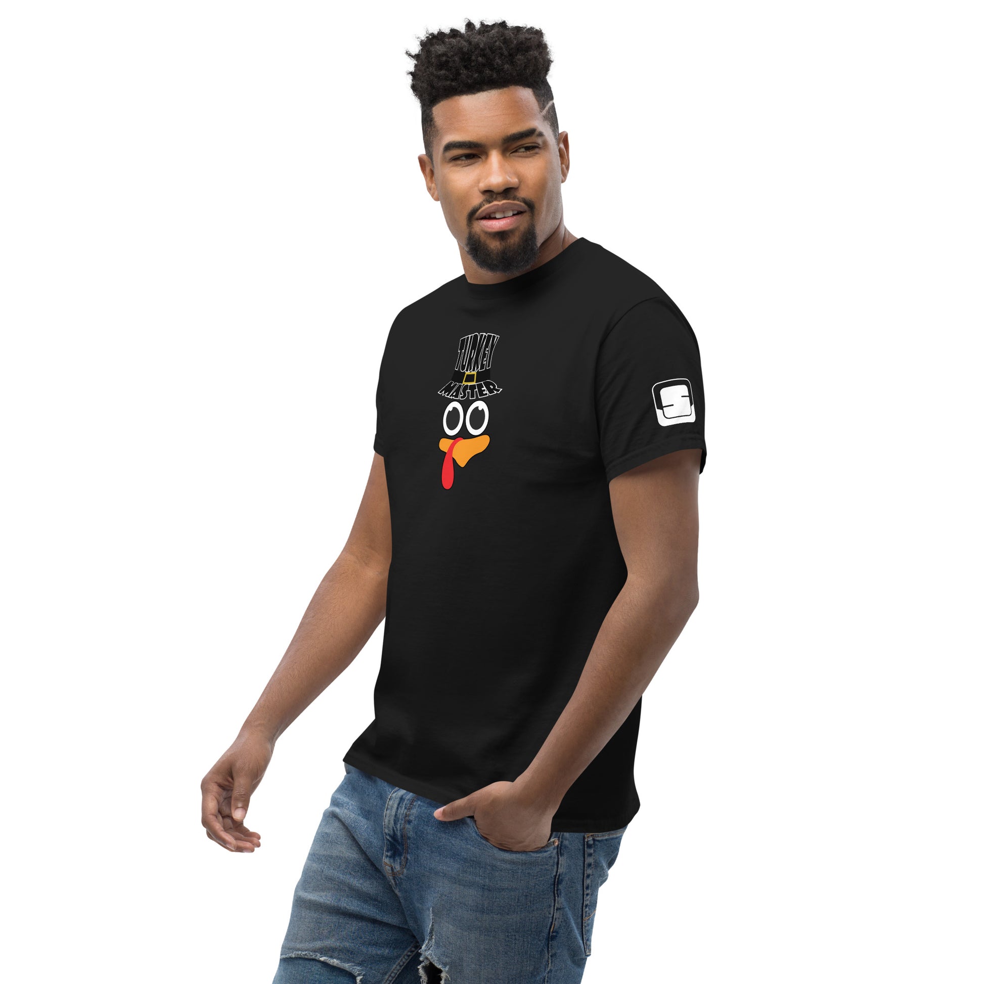 A man with a neatly trimmed beard and curly hairstyle is wearing a black t-shirt featuring a playful turkey graphic. The turkey has cartoonish eyes, an orange beak, and a red snood, along with a black hat that reads "TURKEY MASTER" in bold letters. The left sleeve features a distinctive rectangular logo patch. The man is standing in a casual pose, smiling confidently, with one hand in his pocket. He is wearing distressed jeans, and the background is white.