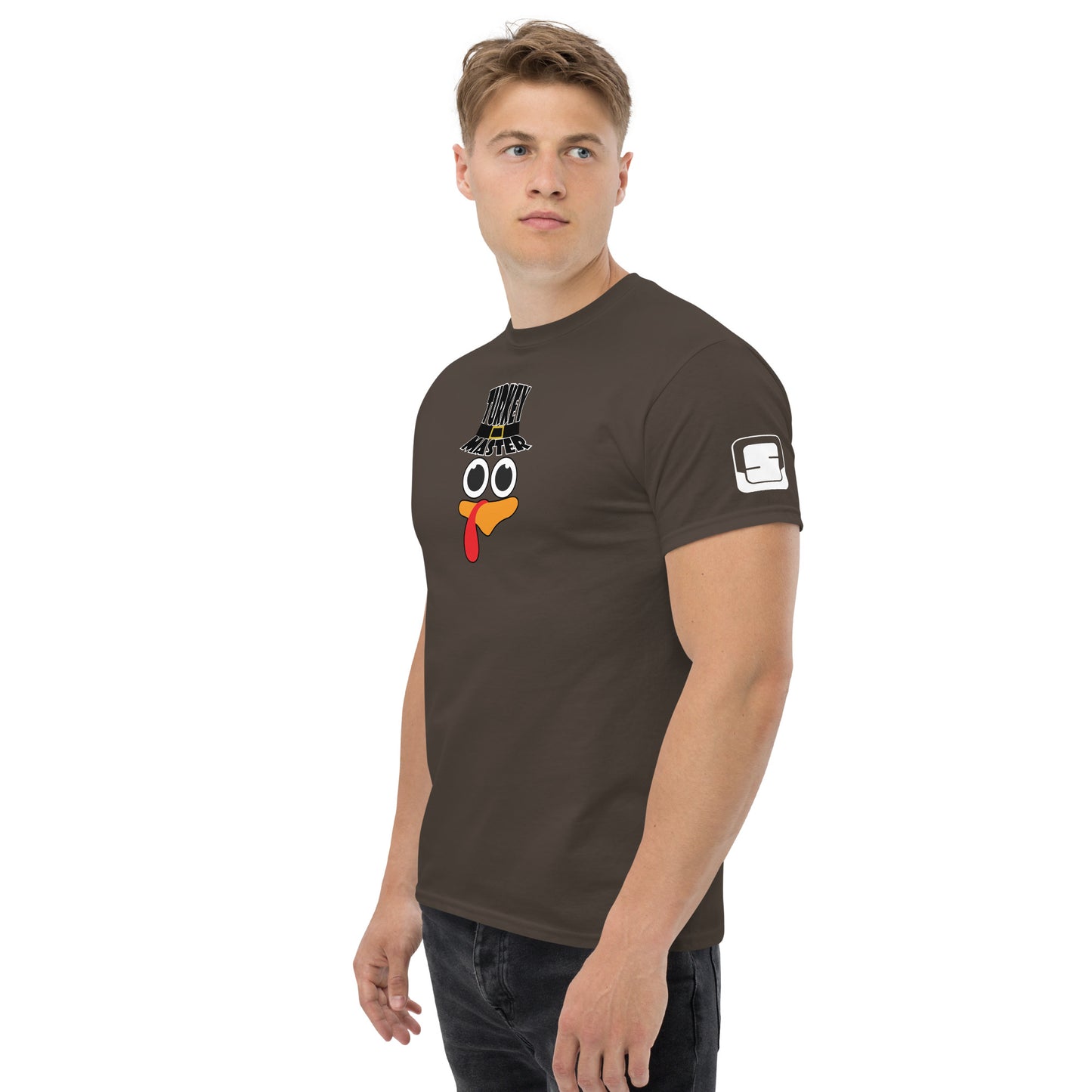 A man with short blonde hair is wearing a brown t-shirt featuring a playful turkey graphic. The turkey has cartoonish eyes, an orange beak, and a red snood, along with a black hat that reads "TURKEY MASTER" in bold letters. On the left sleeve, there's a distinctive rectangular logo patch. The man is standing in a relaxed pose, looking off to the side, against a white background.