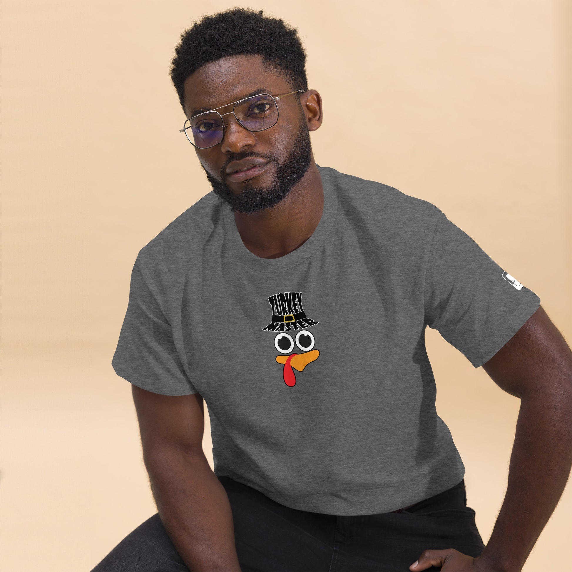 A man with a neatly trimmed beard and glasses is wearing a grey t-shirt featuring a playful turkey graphic. The turkey has cartoonish eyes, an orange beak, and a red snood, along with a black hat reading "TURKEY MASTER" in bold letters. The left sleeve includes a distinctive rectangular logo patch. The man is sitting casually against a neutral background, confidently smiling at the camera.