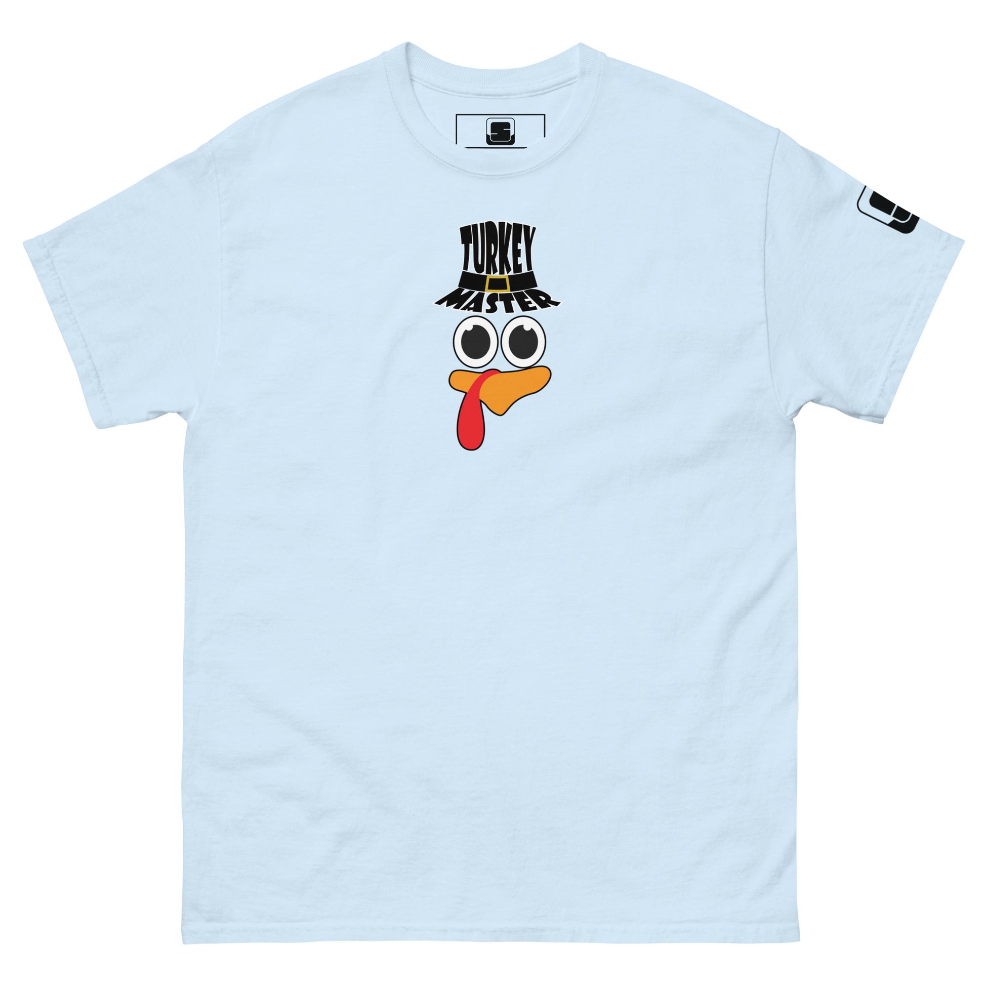 A light-blue t-shirt featuring a playful graphic design of a turkey face with eyes, an orange beak, and a red snood. Above the face, the text reads "TURKEY MASTER" in bold, stylized letters. The text is in the shape of a pilgrims hat. On the left sleeve, there's a rectangular logo patch, adding a unique touch. The shirt is laid out flat, showcasing the entire design clearly.