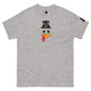 A light grey t-shirt featuring a playful graphic design of a turkey face with eyes, an orange beak, and a red snood. Above the face, the text reads "TURKEY MASTER" in bold, stylized letters. The text is in the shape of a pilgrims hat. On the left sleeve, there's a rectangular logo patch, adding a unique touch. The shirt is laid out flat, showcasing the entire design clearly.