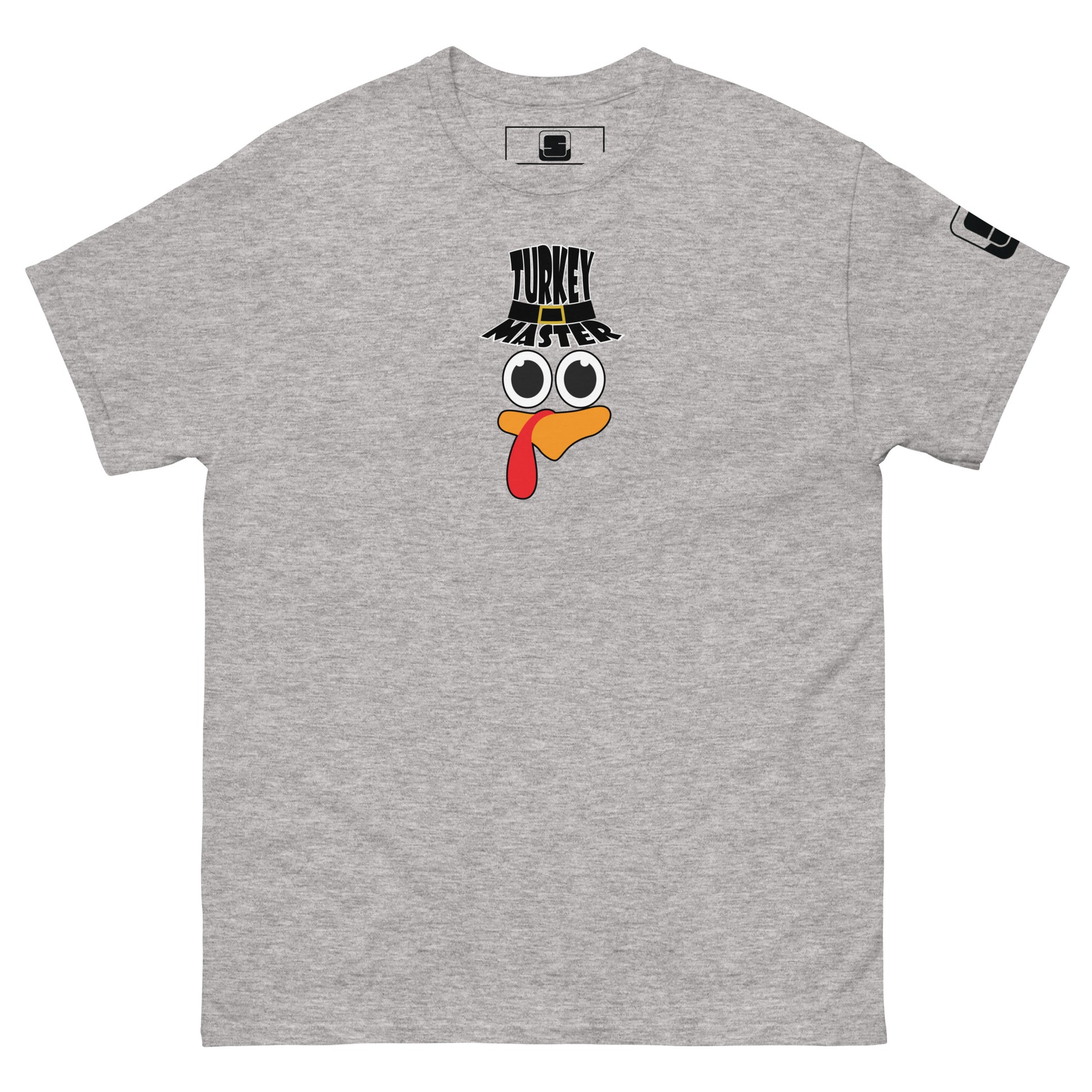 A light grey t-shirt featuring a playful graphic design of a turkey face with eyes, an orange beak, and a red snood. Above the face, the text reads "TURKEY MASTER" in bold, stylized letters. The text is in the shape of a pilgrims hat. On the left sleeve, there's a rectangular logo patch, adding a unique touch. The shirt is laid out flat, showcasing the entire design clearly.