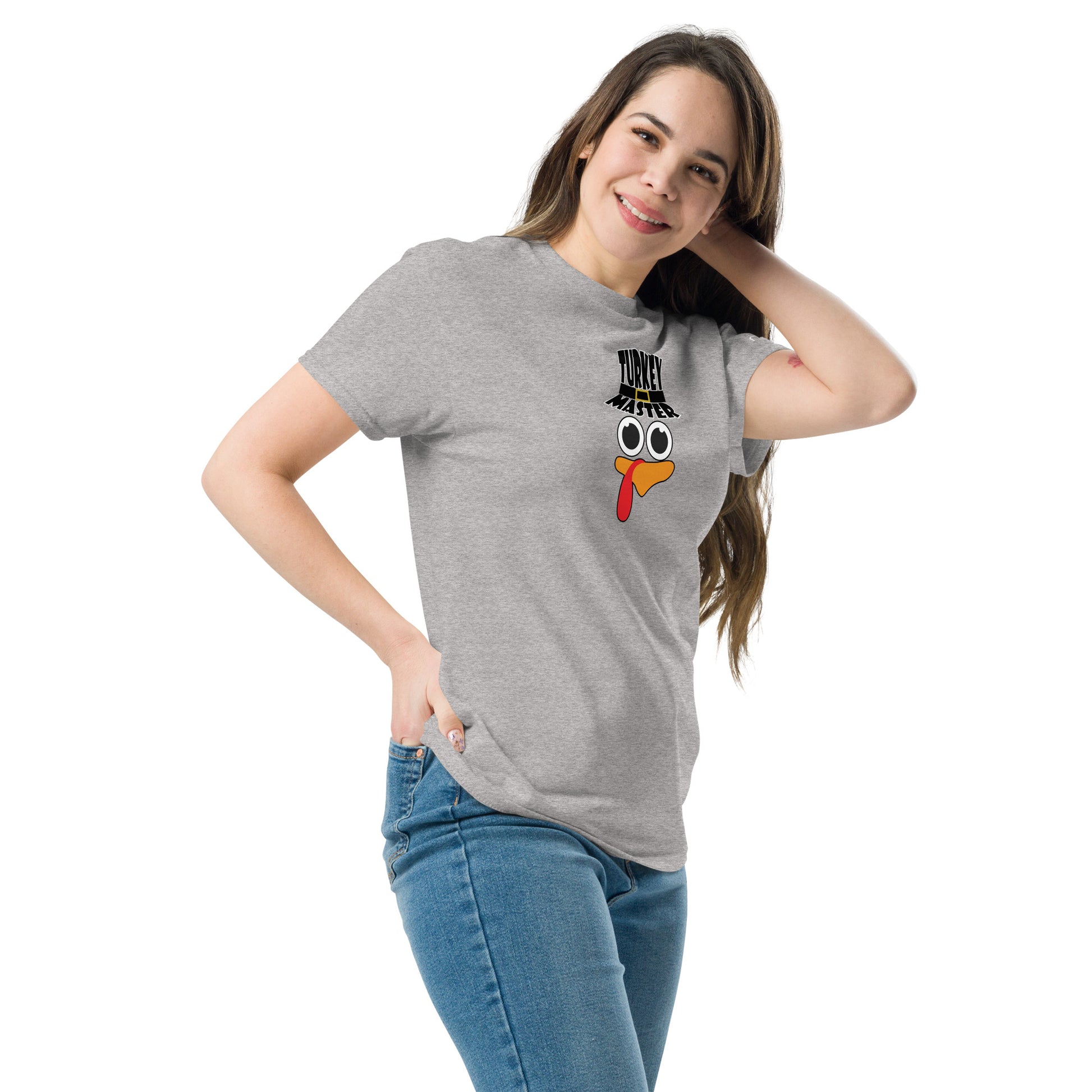 A woman with long brown hair is wearing a grey t-shirt featuring a playful turkey graphic. The turkey has cartoonish eyes, an orange beak, and a red snood, along with a black hat that reads "TURKEY MASTER" in bold letters. She is smiling warmly and posing with one hand on her hip and the other touching her hair, creating a relaxed and confident look. The background is white, highlighting the design of the shirt.