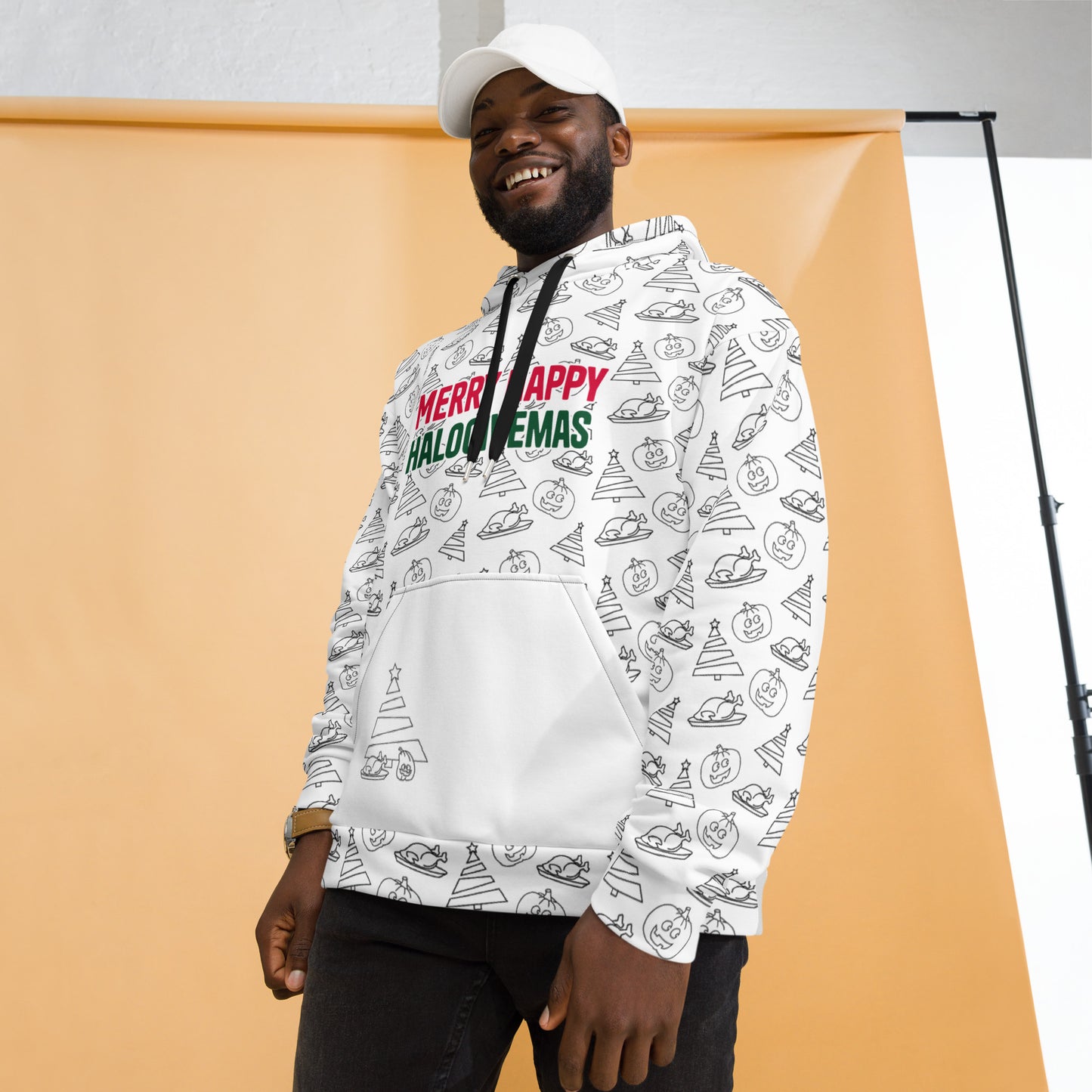 A cheerful man in a white hoodie richly decorated with Halloween, Thanksgiving, and Christmas motifs and the phrase 'Merry Happy Halogivemas', standing against a light background.