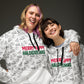 Two women cheerfully posing together in matching white hoodies adorned with a playful print featuring various holiday icons and the phrase 'Merry Happy Halogivemas' in vibrant red and green.
