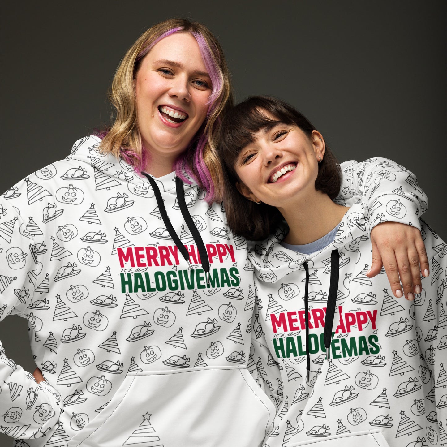 Two women cheerfully posing together in matching white hoodies adorned with a playful print featuring various holiday icons and the phrase 'Merry Happy Halogivemas' in vibrant red and green.