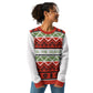 A joyful African American woman with curly hair looks to her side while modeling a festive holiday sweater. The sweater features a colorful array of traditional Christmas patterns in red, green, and white, including geometric diamonds and Christmas trees. A white band with the phrase 'TIS THE SEASON' in green runs across the chest. She has her hands tucked into the pockets of her blue jeans, exuding a relaxed and festive spirit.