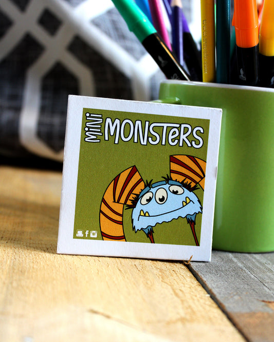 Card with cartoon monster design labeled 'Mini Monsters' on a wooden surface with a green container of pens.