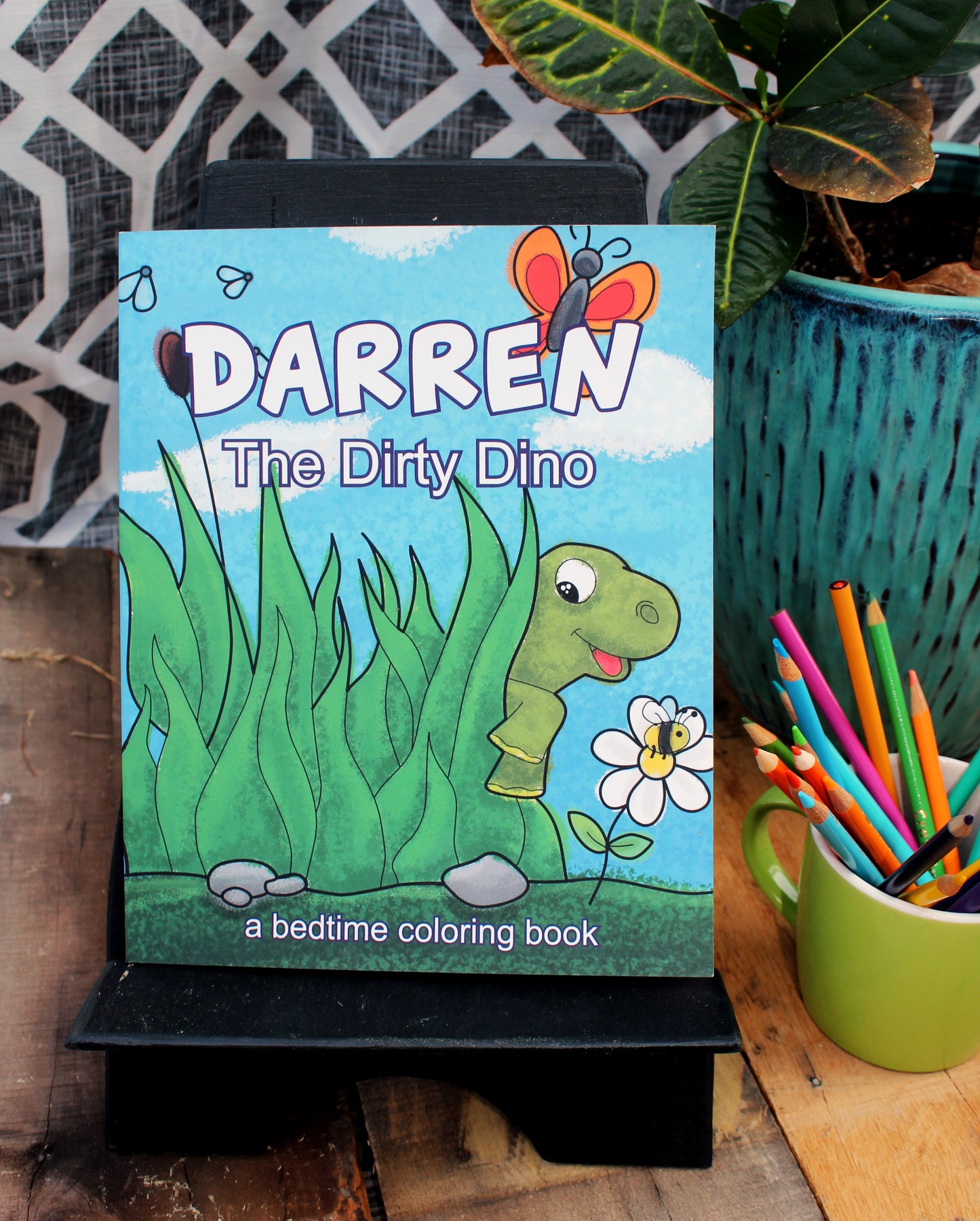 An outdoor display featuring the coloring book 'DARREN The Dirty Dino' on a black easel. The cover shows a cheerful green dinosaur peeking through tall green grass, with a friendly butterfly hovering overhead. Next to the easel is a green mug filled with colorful crayons, set against a backdrop of lush plants and a lattice design.