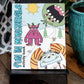 Photograph of the 'Fun in Monsterville Activity Book' propped on a black stand. The vibrant cover features three quirky cartoon monsters: a shy purple monster in the center, a playful blue monster with striped horns at the bottom, and a green monster with an open mouth peering from the top right. The snowy outdoor setting includes pine trees, a winding path, and a bright yellow sun, creating a whimsical scene. A potted plant decorates the right side of the setup.