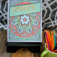 A photo of the "Kolidafun Volume 2" coloring book displayed on a wooden surface with a black stand. The cover features a colorful mandala design with a blue flower at the center, surrounded by geometric patterns. The title, "Kolidafun Volume 2," is written on a diagonal  banner across the top. To the right of the book, a green mug holds an assortment of colorful pencils, enhancing the creative theme of the image. A patterned black-and-white background adds a modern touch.
