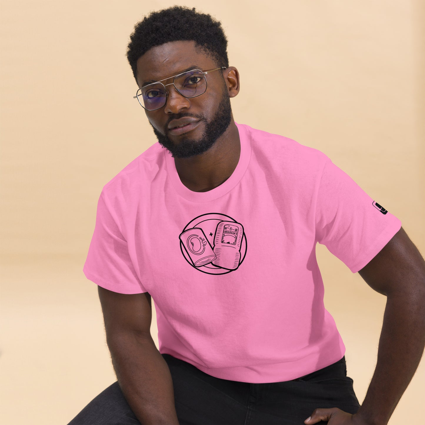  A man sits modeling a bright pink t-shirt featuring a circular emblem in black line art on the chest. The emblem depicts a can of beans and a packet of bologna with a "+" sign between them. He wears glasses and pairs the shirt with dark pants. The t-shirt also includes a small black logo patch on the right sleeve. He poses against a soft beige background, giving the image a relaxed, stylish vibe.
