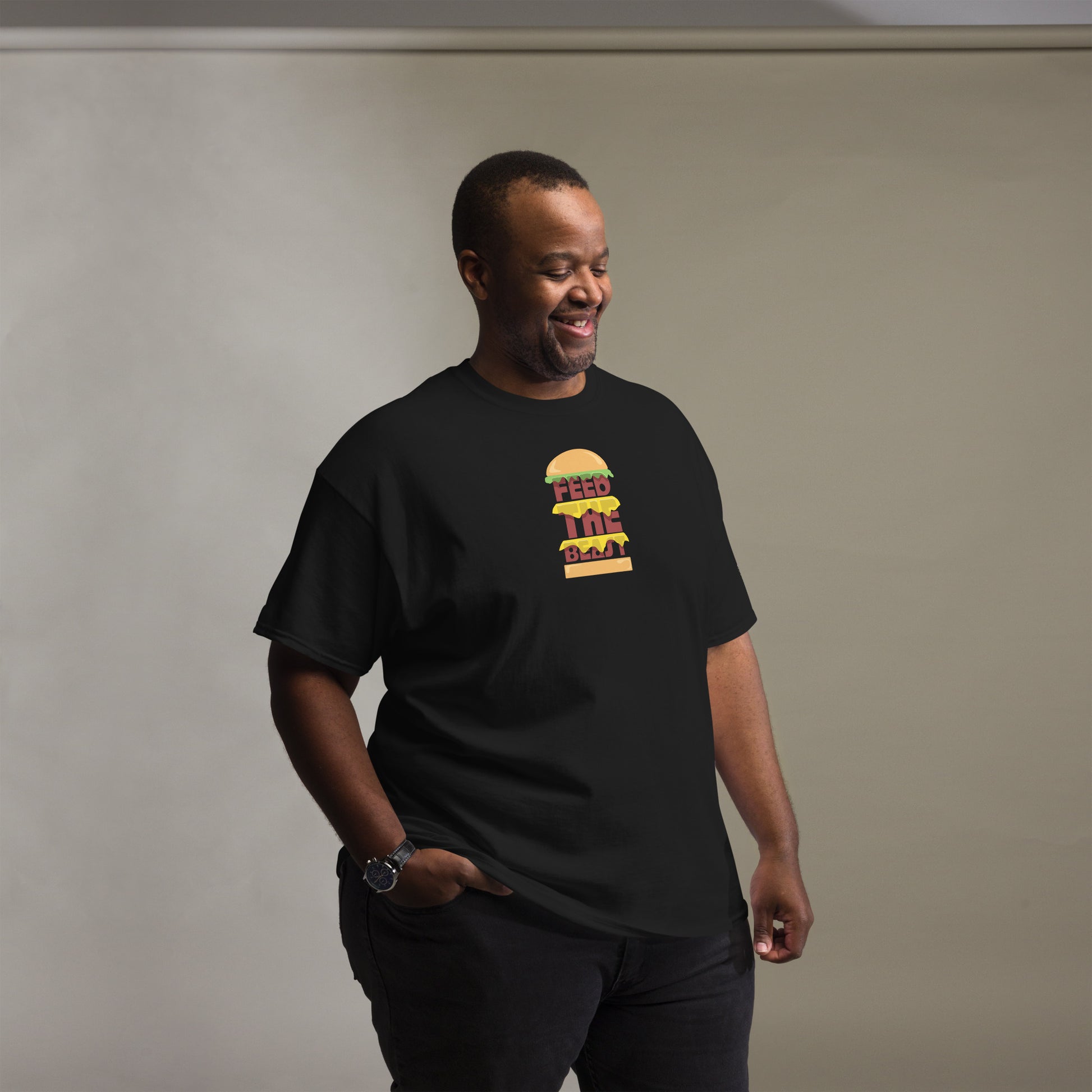 Cheerful man looking down and smiling, wearing a black t-shirt with a colorful 'FEED THE BEAST' burger graphic on the chest, accessorized with a wristwatch, standing in a room with a grey gradient background.