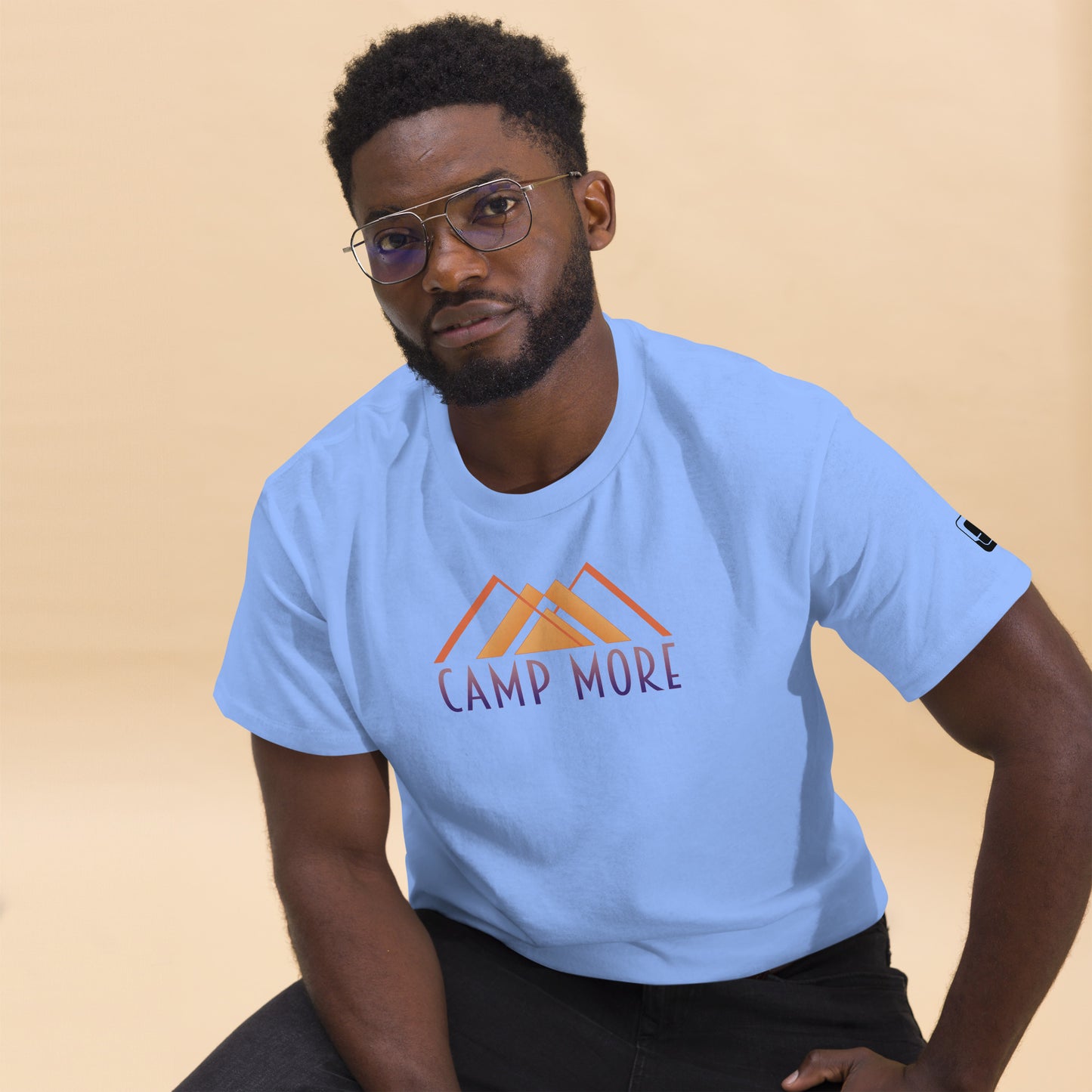 Man wearing glasses looking confidently at the camera, donned in a light blue t-shirt with the phrase 'CAMP MORE' in stylized orange lettering below a minimalist mountain graphic, complete with a logo patch on the sleeve, set against a soft beige background.