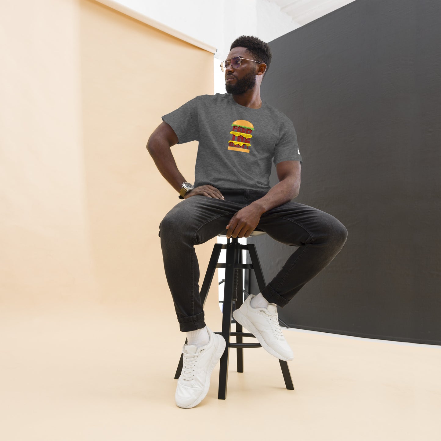 Stylish man with a beard and glasses, wearing a dark gray t-shirt with a colorful stacked burger graphic and 'FEED THE BEAST' text, seated on a tall stool, sporting black jeans and white sneakers, against a peach and black corner wall background.