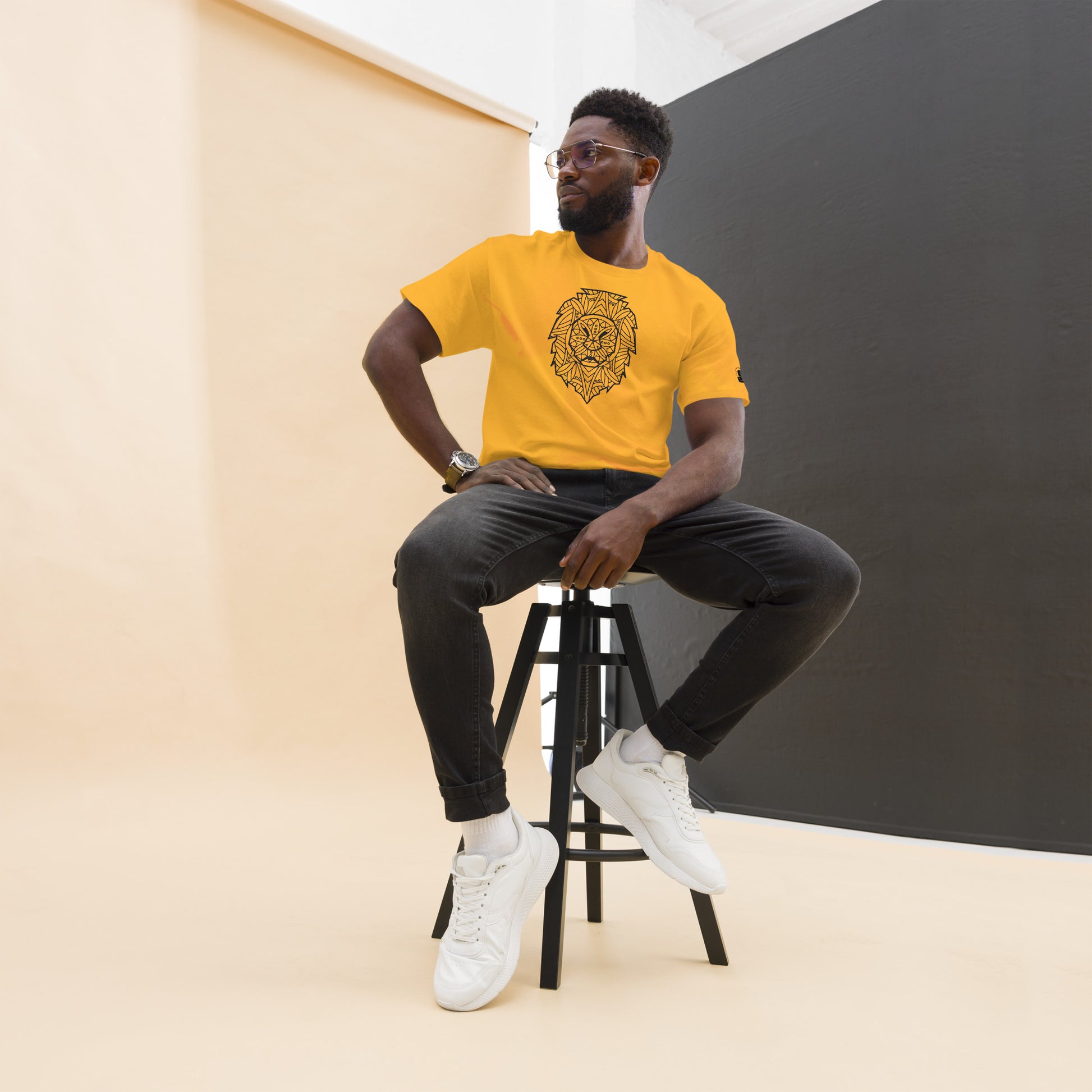 Stylish man in glasses perched on a stool, sporting a mustard yellow t-shirt with a bold black lion head graphic, paired with dark jeans and white sneakers, in a room with contrasting beige and black walls.