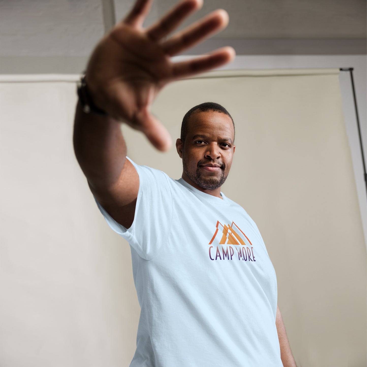 Man in a light blue t-shirt with  in stylized orange lettering below a minimalist mountain graphic reaching out towards the camera in a stopping gesture, giving a serious expression, against a beige backdrop.