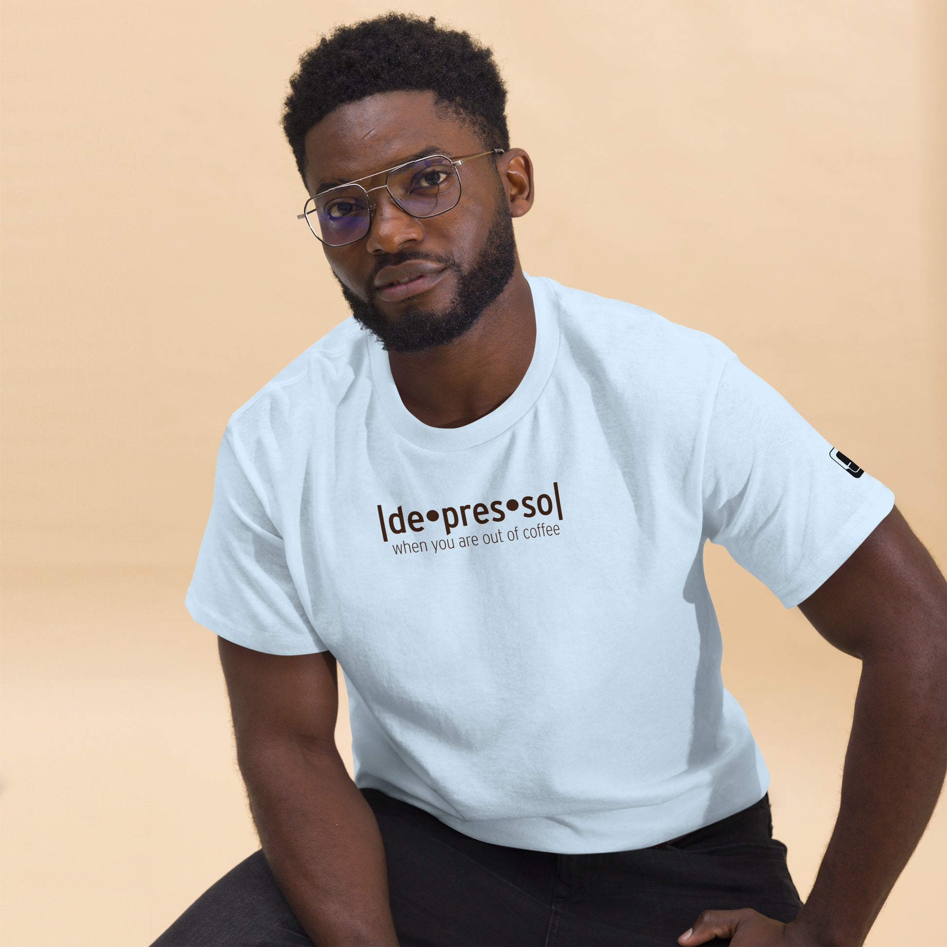 Man with glasses looking to the side, exuding a contemplative vibe, dressed in a light blue t-shirt with the witty phrase 'depresso' followed by 'when you are out of coffee', along with a logo patch on the sleeve, set against a soft beige background.