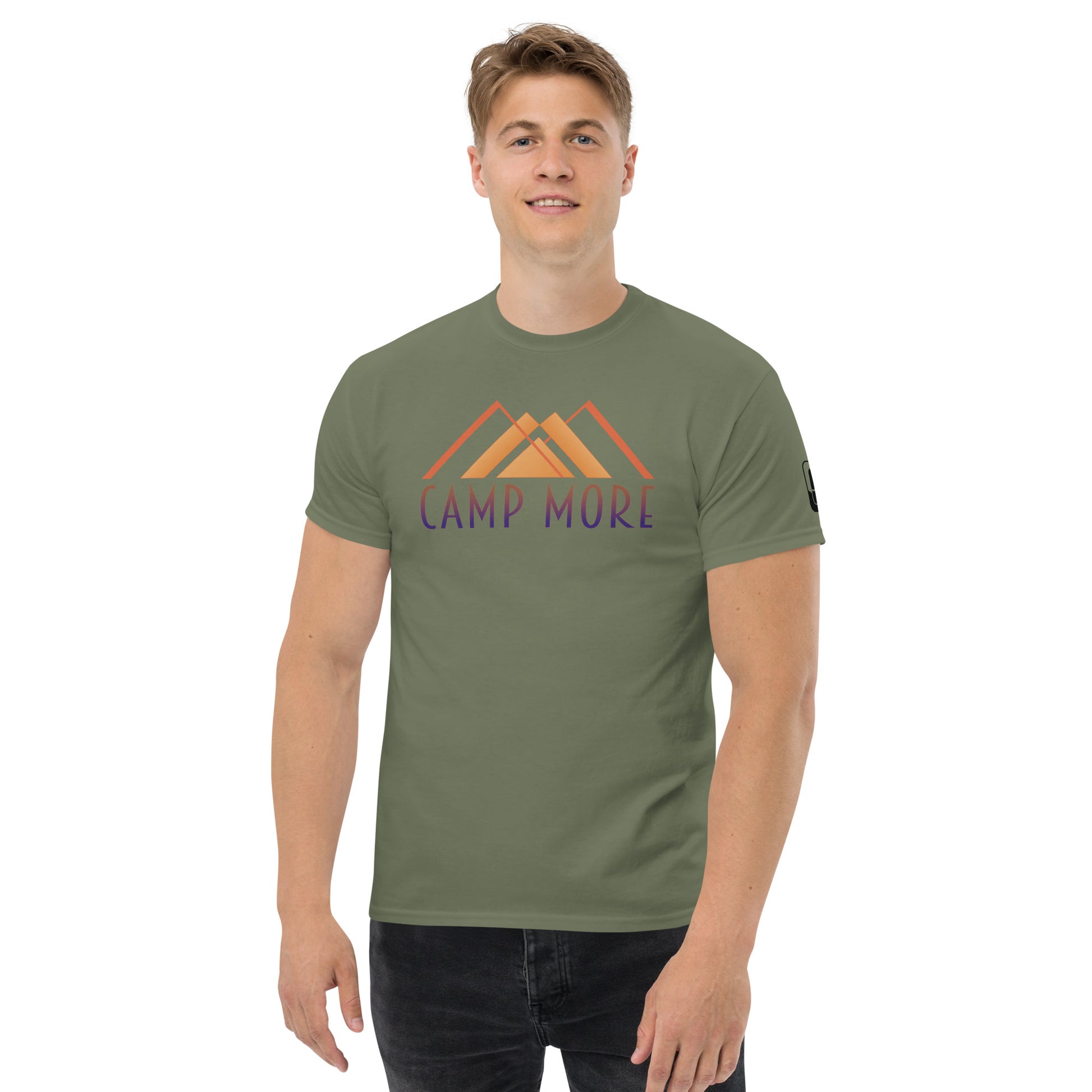 Smiling young man wearing an military green t-shirt with the 'CAMP MORE' slogan in stylized lettering over a simple mountain graphic, and a logo patch on the sleeve, against a white background