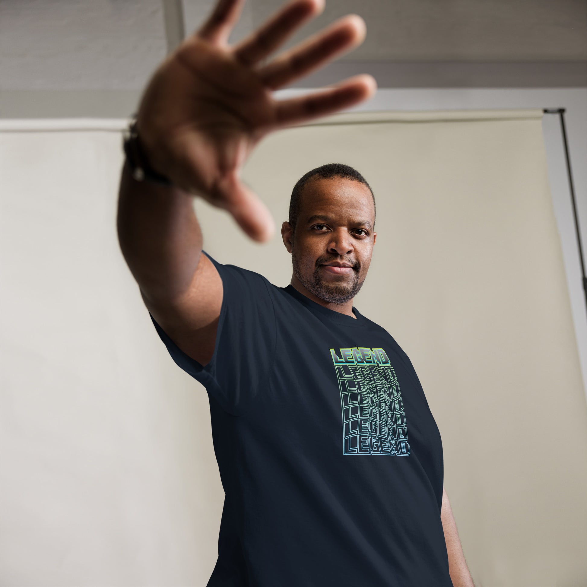 Man extending his hand towards the camera, offering a friendly yet assertive gesture, wearing a navy t-shirt with a 'LEGEND' cube graphic in a green-to-light blue gradient, complemented by a watch and set against a light grey backdrop.