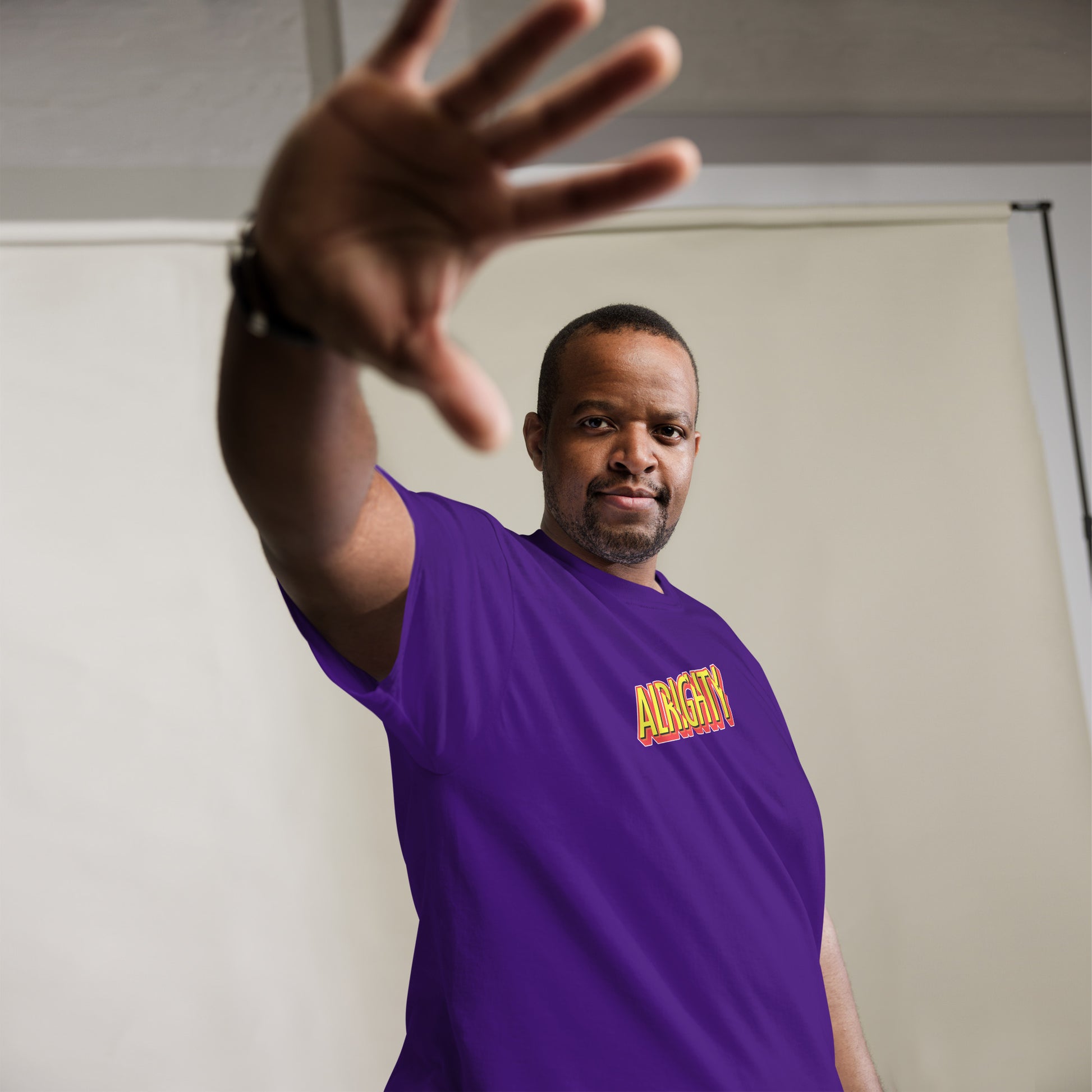 Man extending his hand towards the camera with a firm expression, wearing a vibrant purple t-shirt emblazoned with the word 'ALRIGHTY' in a fiery font, against a white backdrop with soft shadows.