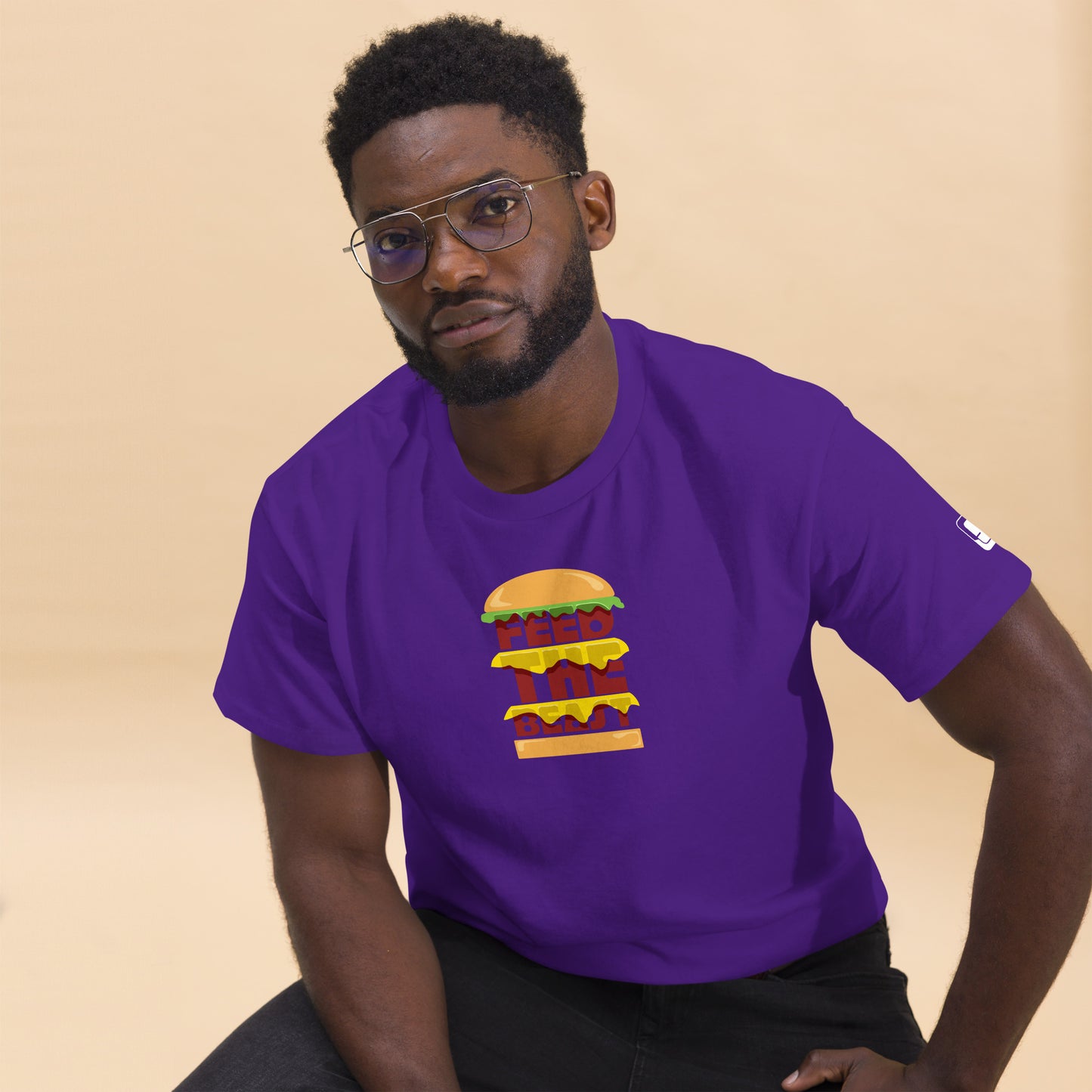 Man wearing glasses and looking thoughtfully to the side, clad in a vibrant purple t-shirt with a colorful burger graphic and the words 'FEED THE BEAST' in yellow, seated against a warm beige backdrop.