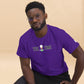  A man wearing glasses poses in a vibrant purple t-shirt with a "TEE TIME" logo featuring a golf ball and tee. The design is placed centrally on the chest. His expression is thoughtful, and he's looking directly at the viewer. A small logo is visible on the sleeve. He pairs the shirt with black pants against a soft beige background, giving the image a warm, approachable feel while highlighting the bold shirt color and crisp design.