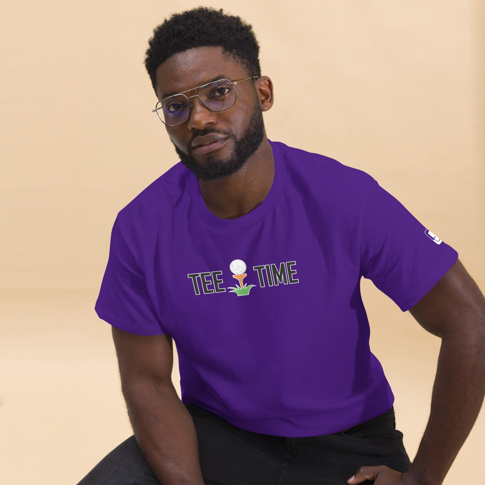  A man wearing glasses poses in a vibrant purple t-shirt with a "TEE TIME" logo featuring a golf ball and tee. The design is placed centrally on the chest. His expression is thoughtful, and he's looking directly at the viewer. A small logo is visible on the sleeve. He pairs the shirt with black pants against a soft beige background, giving the image a warm, approachable feel while highlighting the bold shirt color and crisp design.