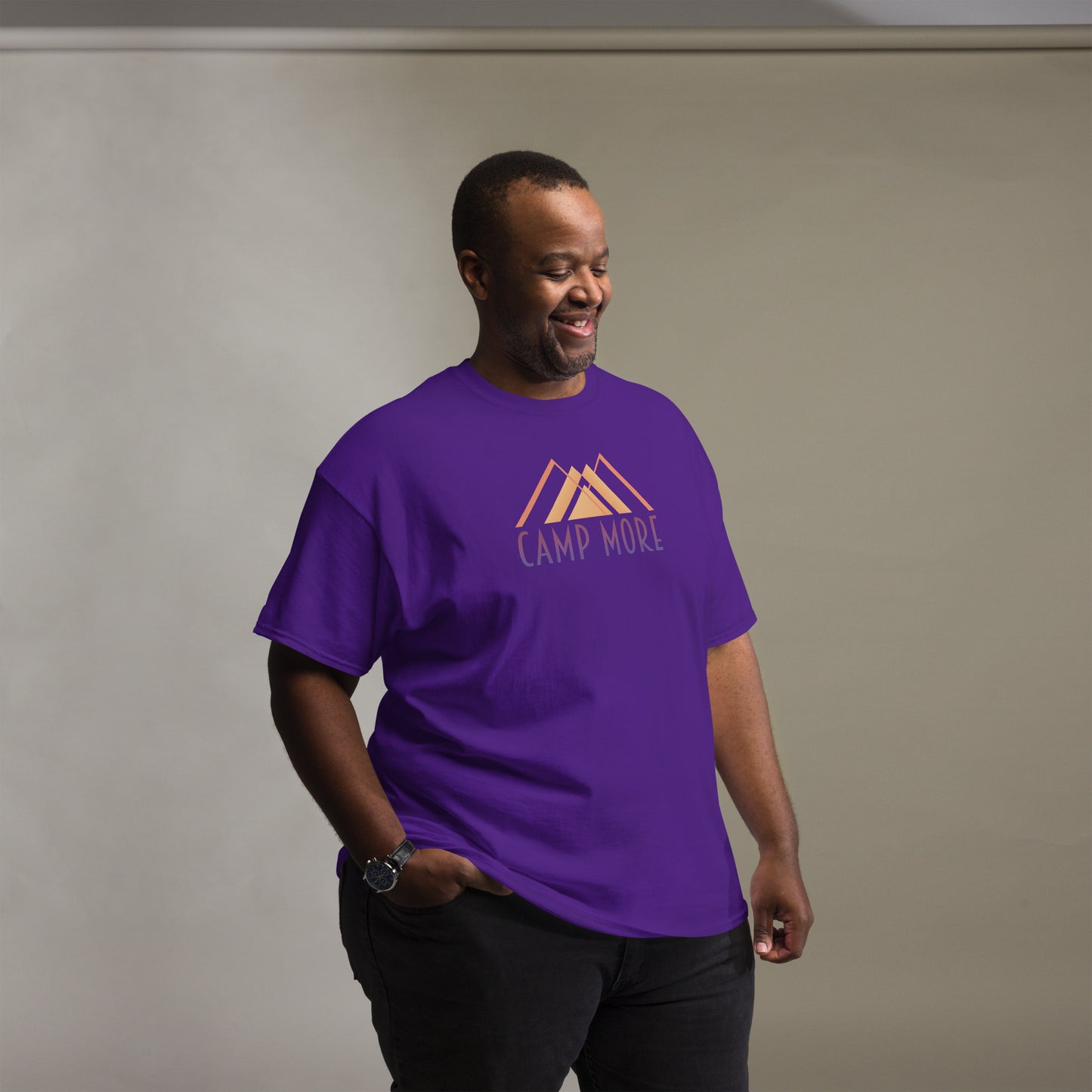 Cheerful man wearing a purple t-shirt with the orange 'CAMP MORE' slogan and abstract mountain design, complemented with black pants and a watch, standing in a room with a grey background and soft lighting