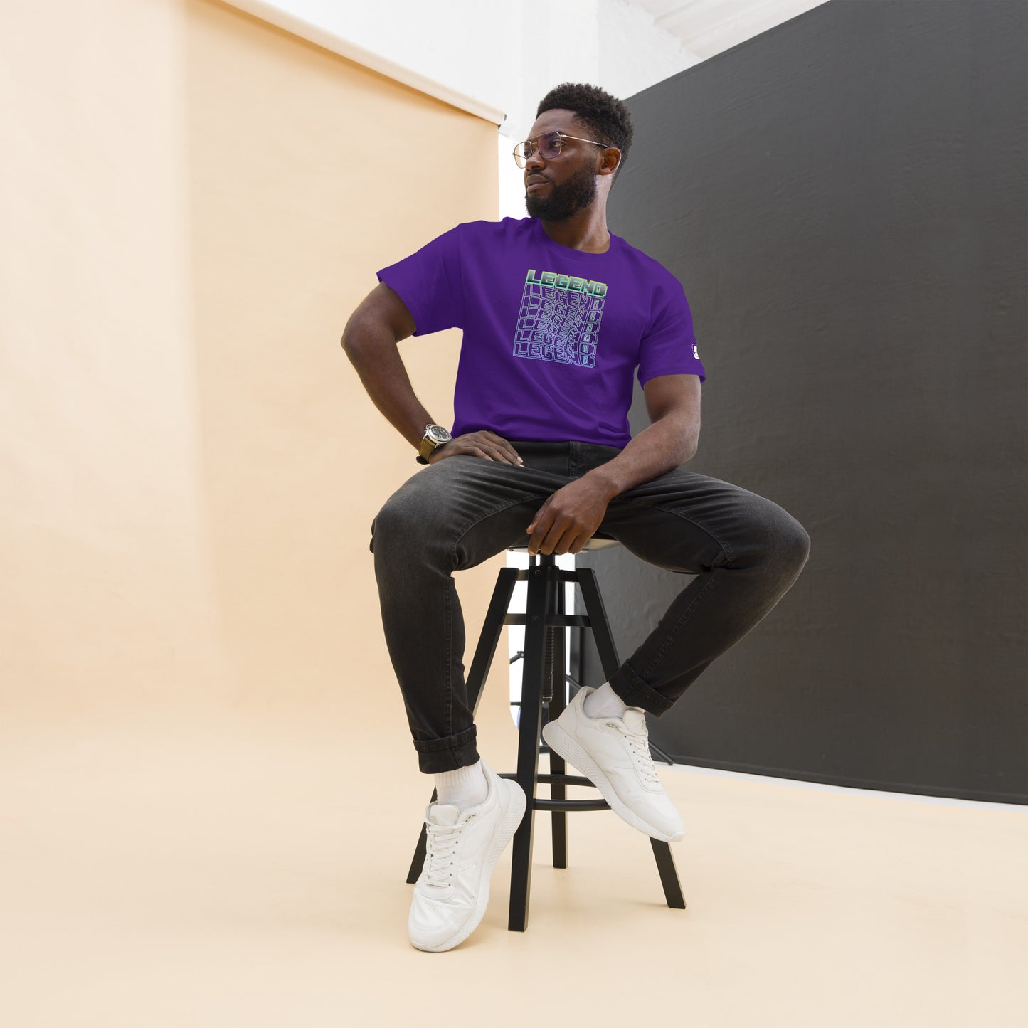 Stylish man seated on a high stool, looking away thoughtfully, wearing a purple t-shirt with a cube-shaped 'LEGEND' graphic in a green-to-light blue gradient, completed with dark jeans, white sneakers, and a wristwatch, against a two-tone peach and black background.