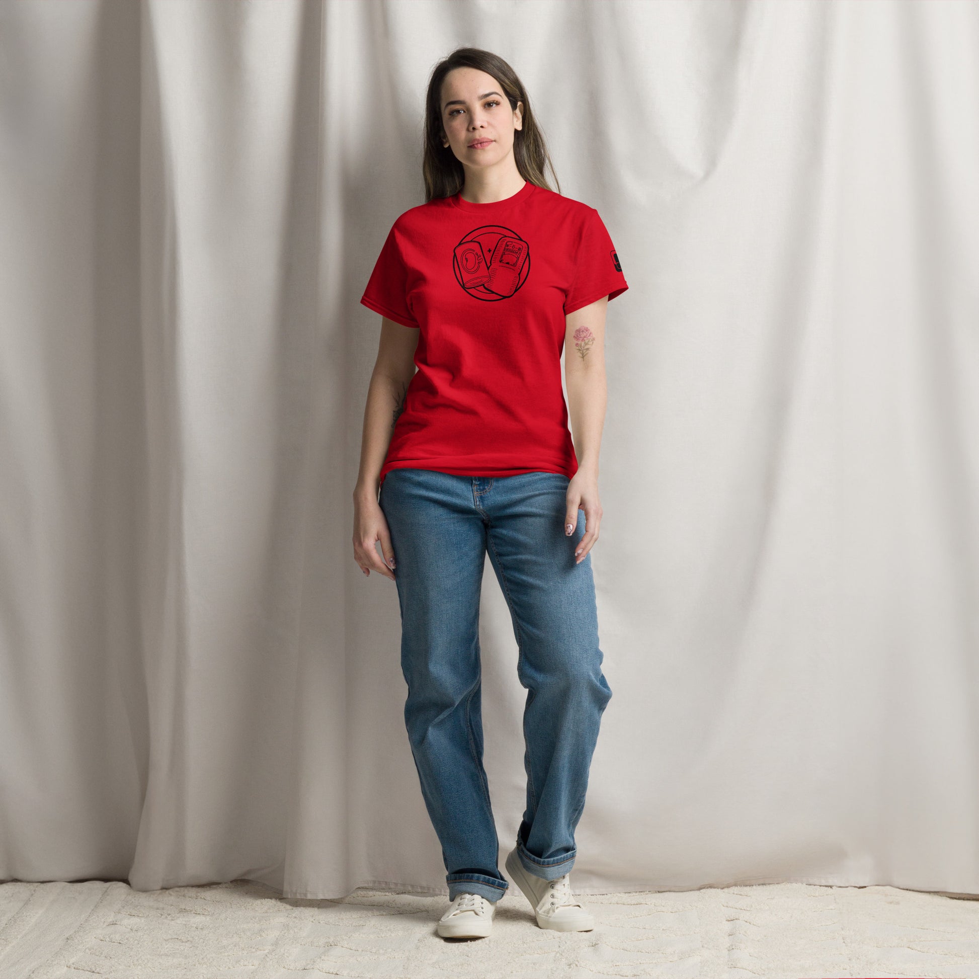 A woman stands confidently in a red t-shirt featuring a circular emblem in black line art, which includes a can of beans and some bologna with a "+" sign between them. She pairs the shirt with blue jeans and white sneakers. The t-shirt also has a small logo patch on the right sleeve. She poses against a neutral draped fabric background, giving the image a relaxed, casual vibe that emphasizes the shirt's playful and stylish design.