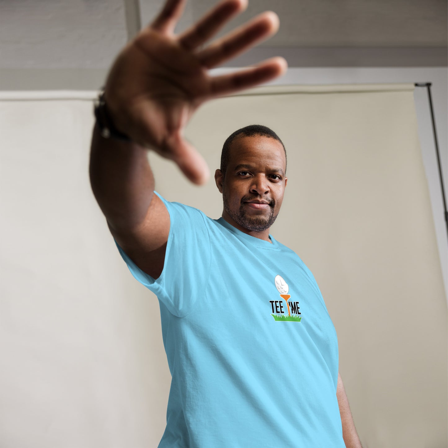 Man wearing a light blue short-sleeved t-shirt with a 'Tee Time' graphic showing a golf ball character on grass on the chest standing against a plain beige backdrop with an out stitched arm.
