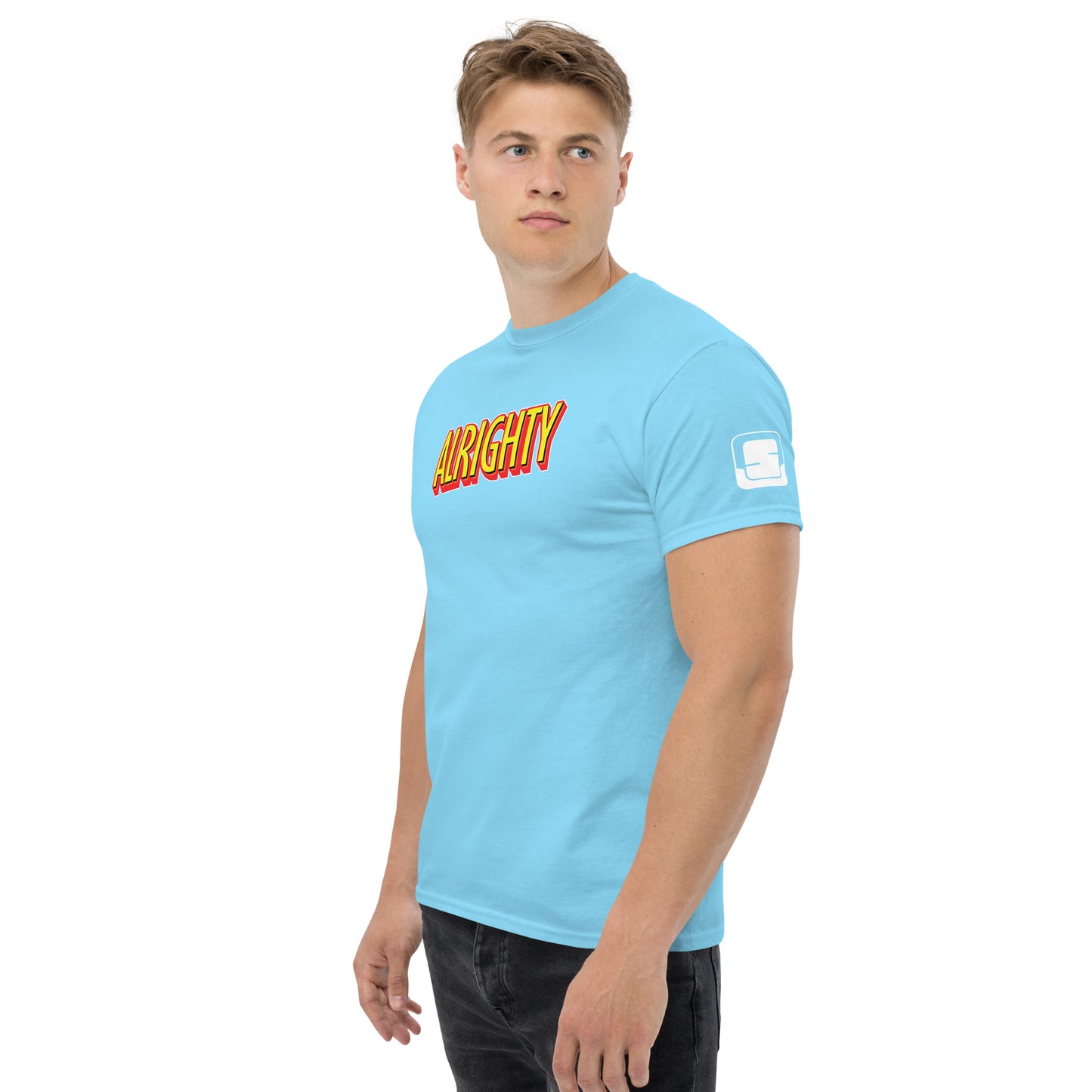 Focused young man in a sky blue t-shirt with the word 'ALRIGHTY' in bold, flame-like font across the chest, a logo patch on the sleeve, paired with dark jeans, standing against a neutral backdrop.