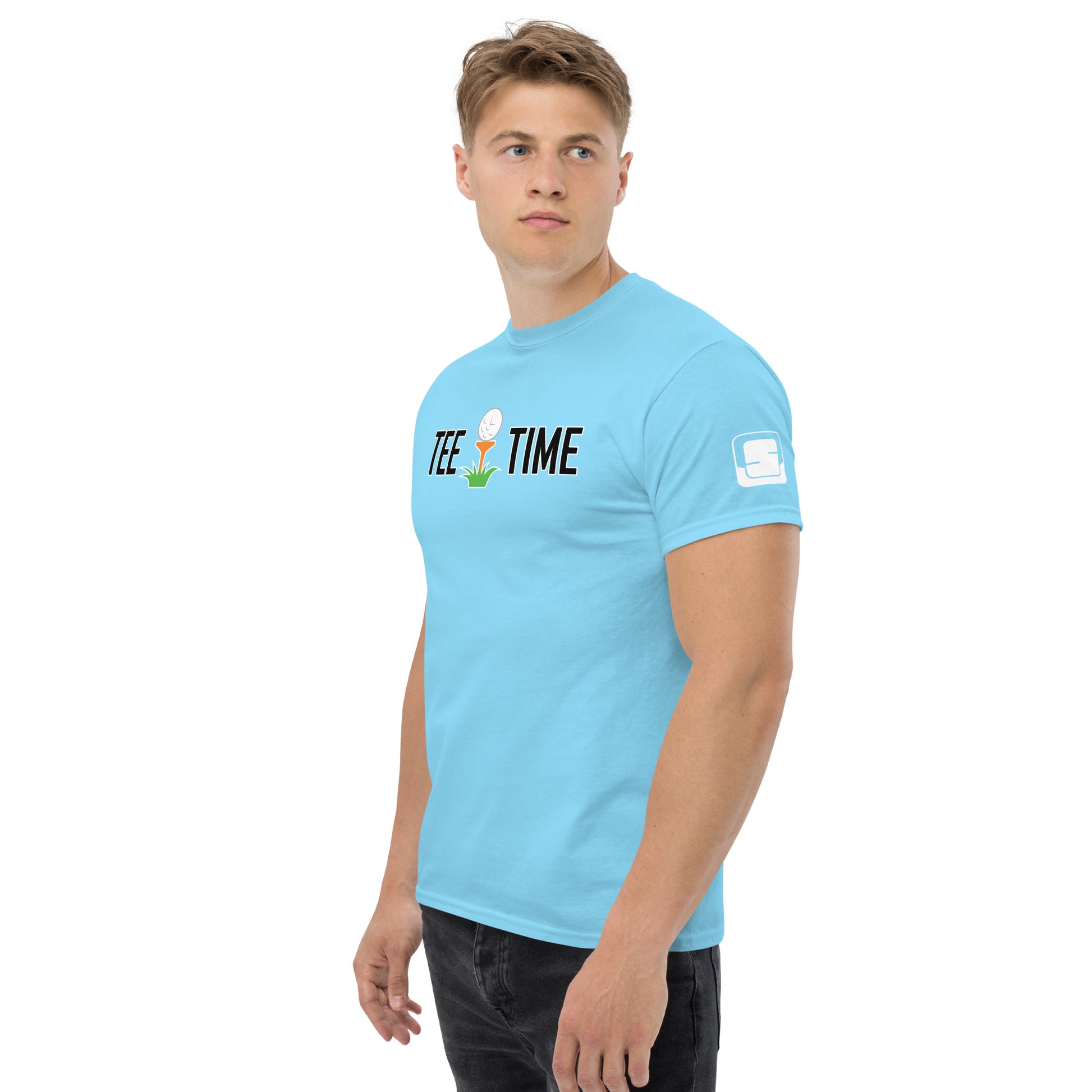 A young man models a light blue t-shirt featuring the phrase "TEE TIME" with an illustration of a golf ball on a tee above grass. He looks off to the side with a focused expression. The shirt has a small square logo on the sleeve. His hairstyle is neat, and he wears dark pants, complementing the casual yet stylish tee. The background is neutral, emphasizing the shirt's design and the model's clean-cut presentation.