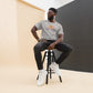 Man with glasses seated casually on a black stool, wearing a heather grey 'CAMP MORE' t-shirt with an orange and an abstract orange mountain design, black jeans, white sneakers, and a wristwatch, in front of a contrasting black and cream backdrop.
