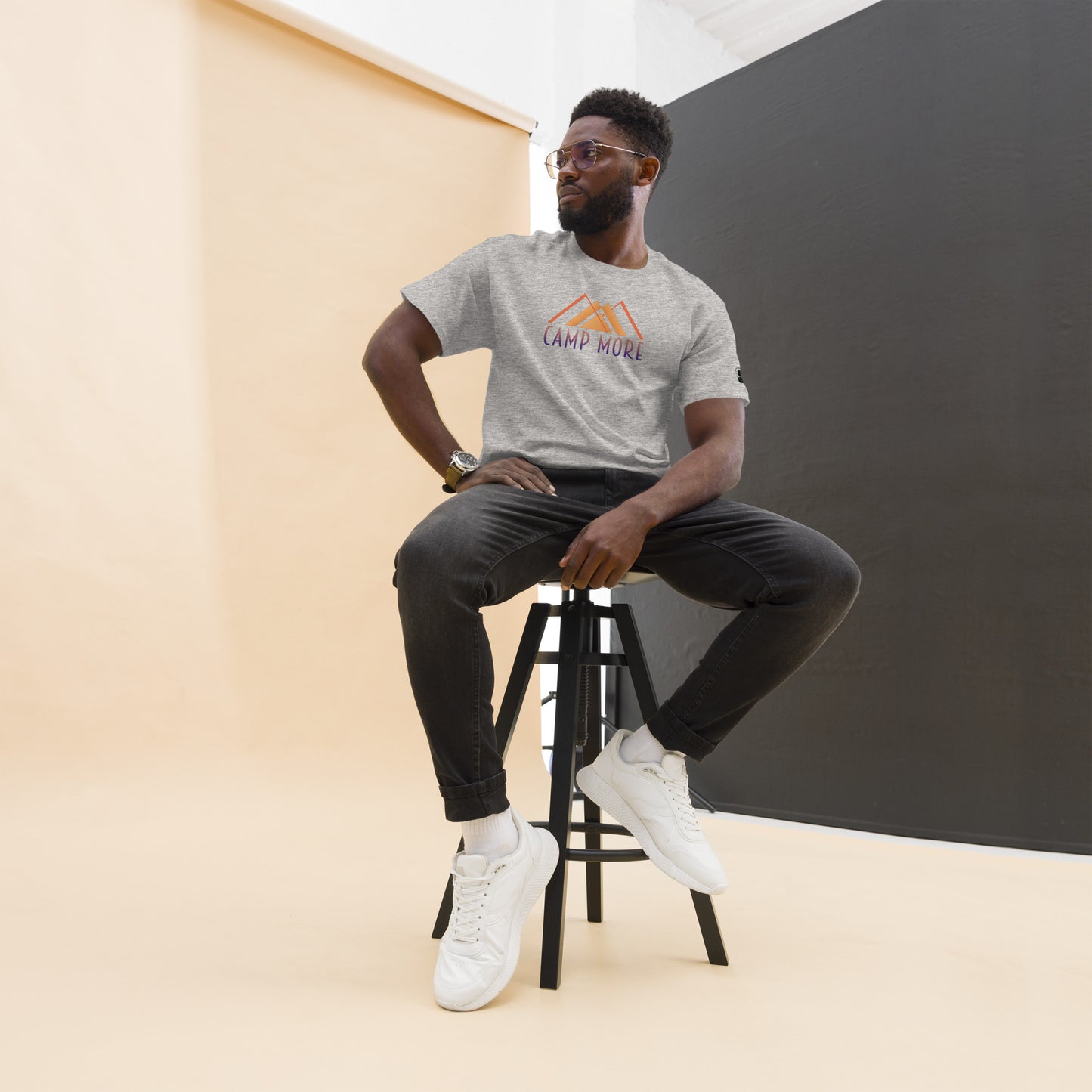 Man with glasses seated casually on a black stool, wearing a heather grey 'CAMP MORE' t-shirt with an orange and an abstract orange mountain design, black jeans, white sneakers, and a wristwatch, in front of a contrasting black and cream backdrop.