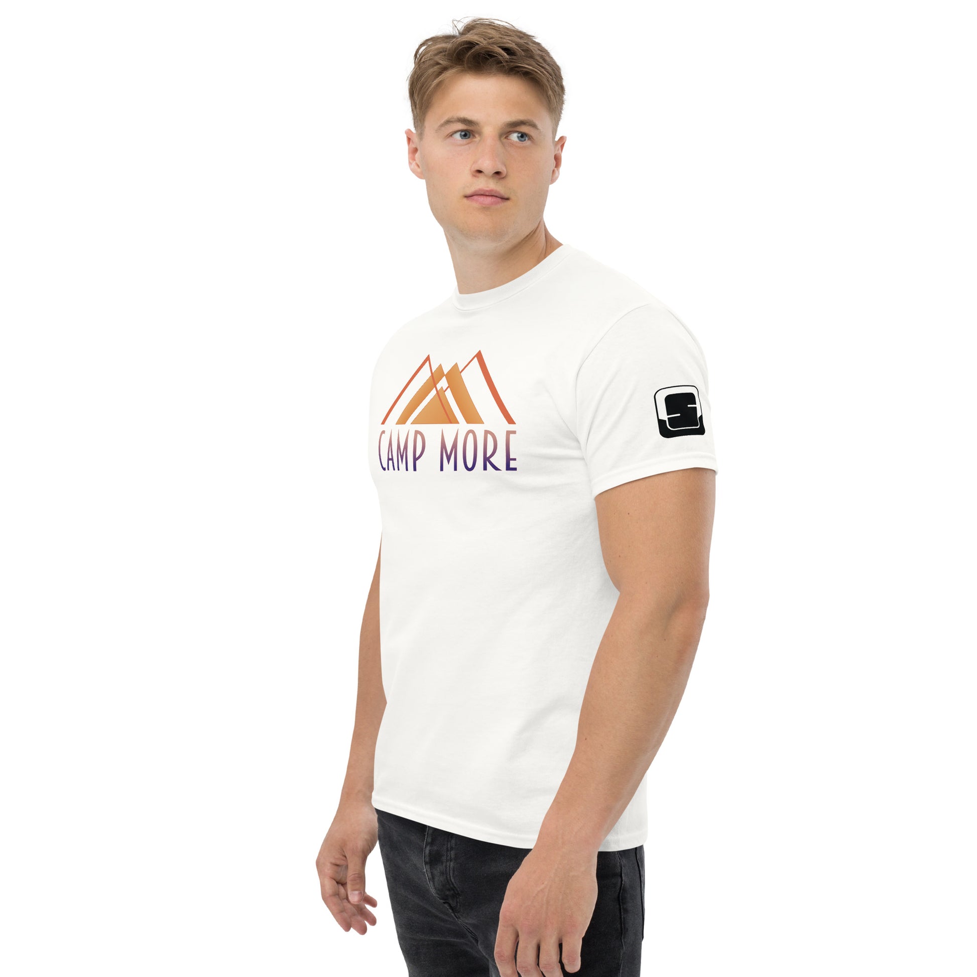 Confident young man in a white t-shirt with 'CAMP MORE' written above an abstrict mountain graphic in orange and purple. There is also a small square logo patch on the sleeve. This shirt is paired with dark trousers, against a white background.