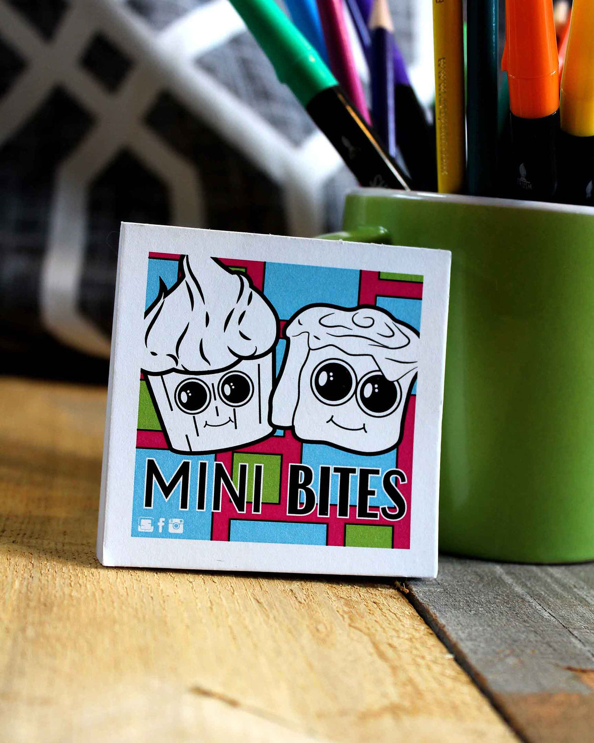 Sticker with cartoon ice cream characters and 'Mini Bites' text on a wooden surface with a green container of pens.