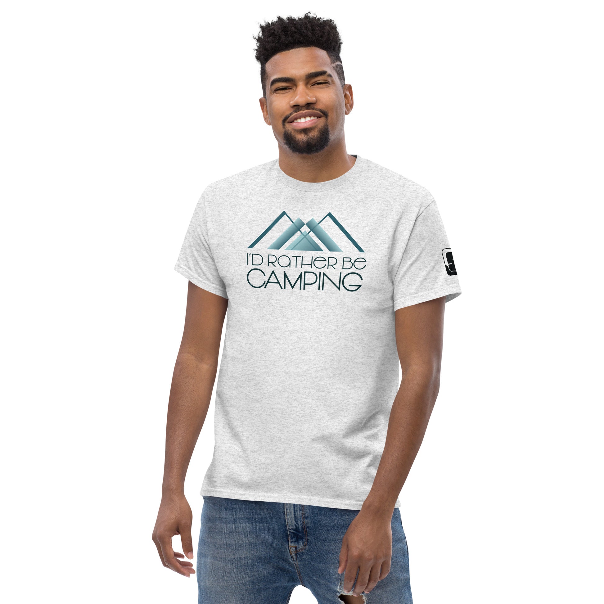 A young African American man with a friendly smile, wearing a light grey t-shirt that reads "I'D RATHER BE CAMPING" with a stylized teal mountain graphic above the text. He stands relaxed, with his left hand slightly in front of his denim jeans, against a white background. His casual, carefree demeanor and stylish haircut complement the outdoor-themed shirt he's modeling.