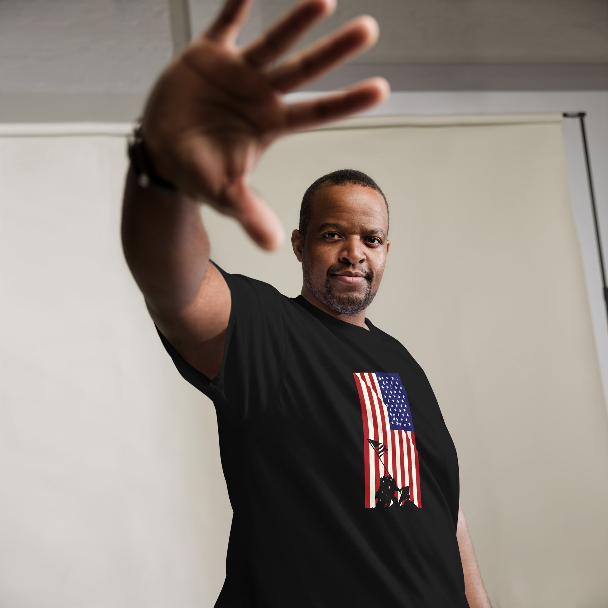 A confident man reaches towards the camera in a gesture that blends invitation and assertion. He sports a black T-shirt featuring a bold graphic of the American flag, behind which the iconic Iwo Jima silhouette is prominently displayed. The combination of his assertive pose and the patriotic design creates a powerful statement, set against a simple studio backdrop that keeps the focus on him and his attire.