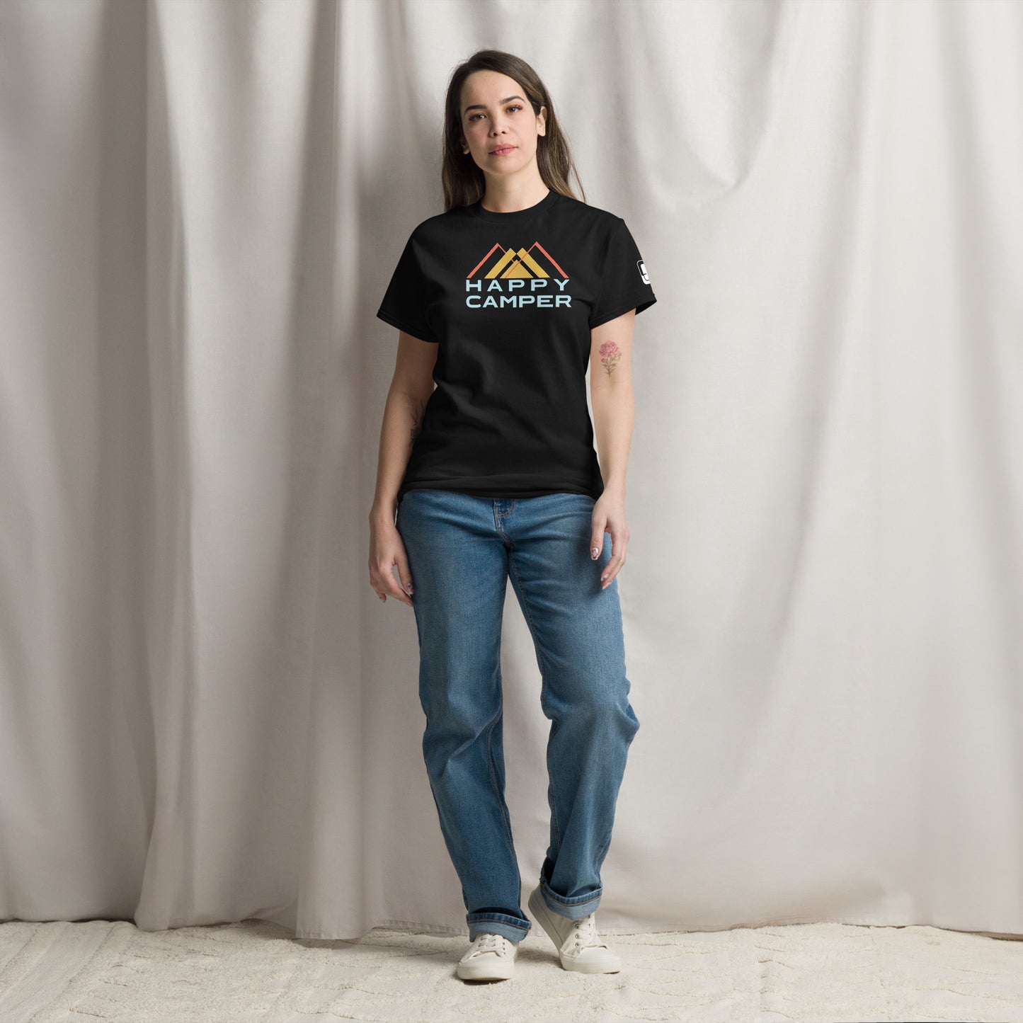 A woman stands confidently in a studio setting, wearing a black t-shirt emblazoned with the phrase "HAPPY CAMPER" in bold, colorful letters below a stylized mountain graphic. She is casually dressed in loose-fitting blue jeans and white sneakers, with her dark hair pulled back. The simple background highlights her relaxed, poised demeanor.
