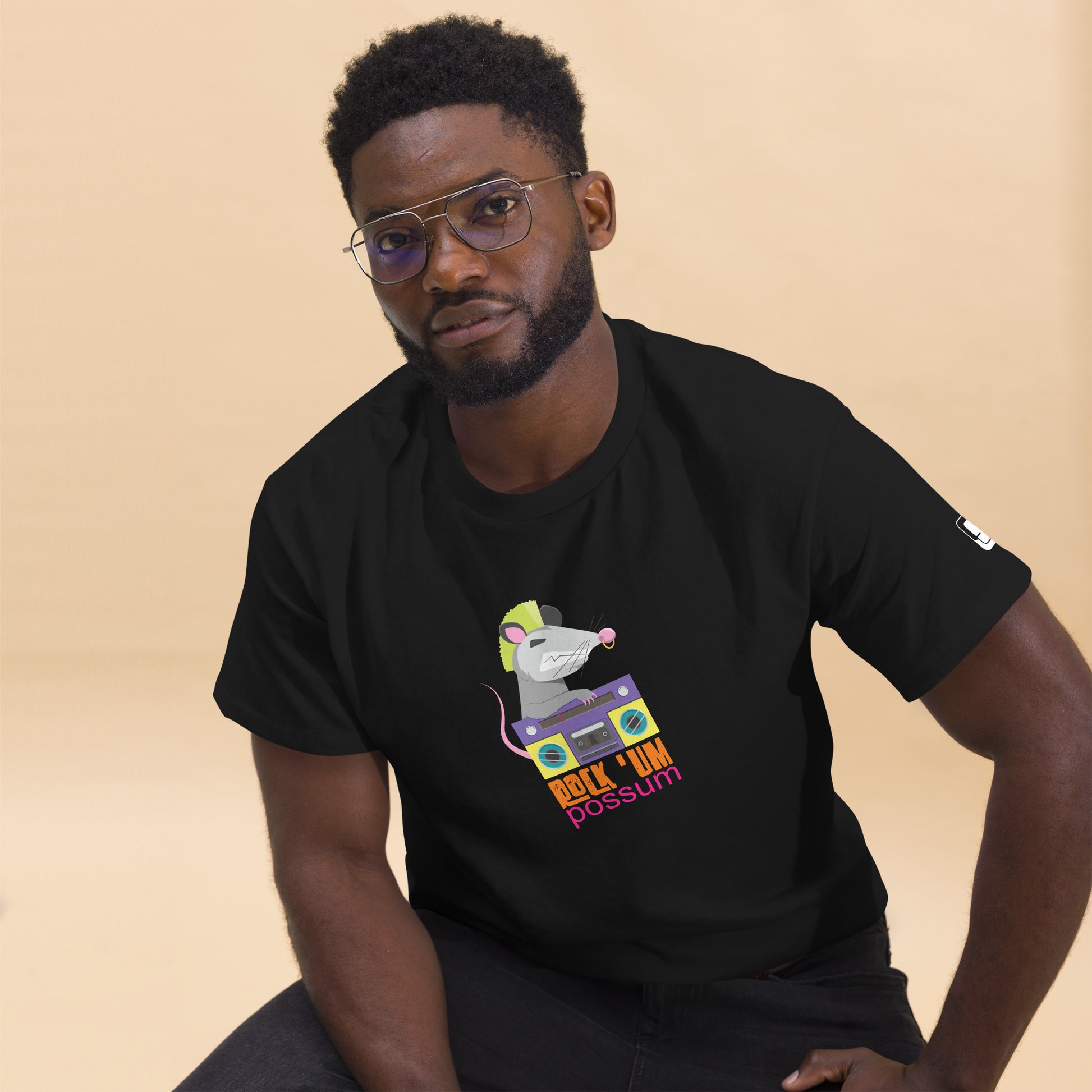 A confident African American man with a neatly trimmed beard and stylish glasses, posing while seated. He wears a sleek black t-shirt adorned with a whimsical graphic of a possum sporting a neon green mohawk and handling a colorful purple boombox. The shirt features the playful text "ROCK 'UM POSSUM" below the image. His casual yet thoughtful expression and modern style enhance the shirt's funky and youthful appeal.

