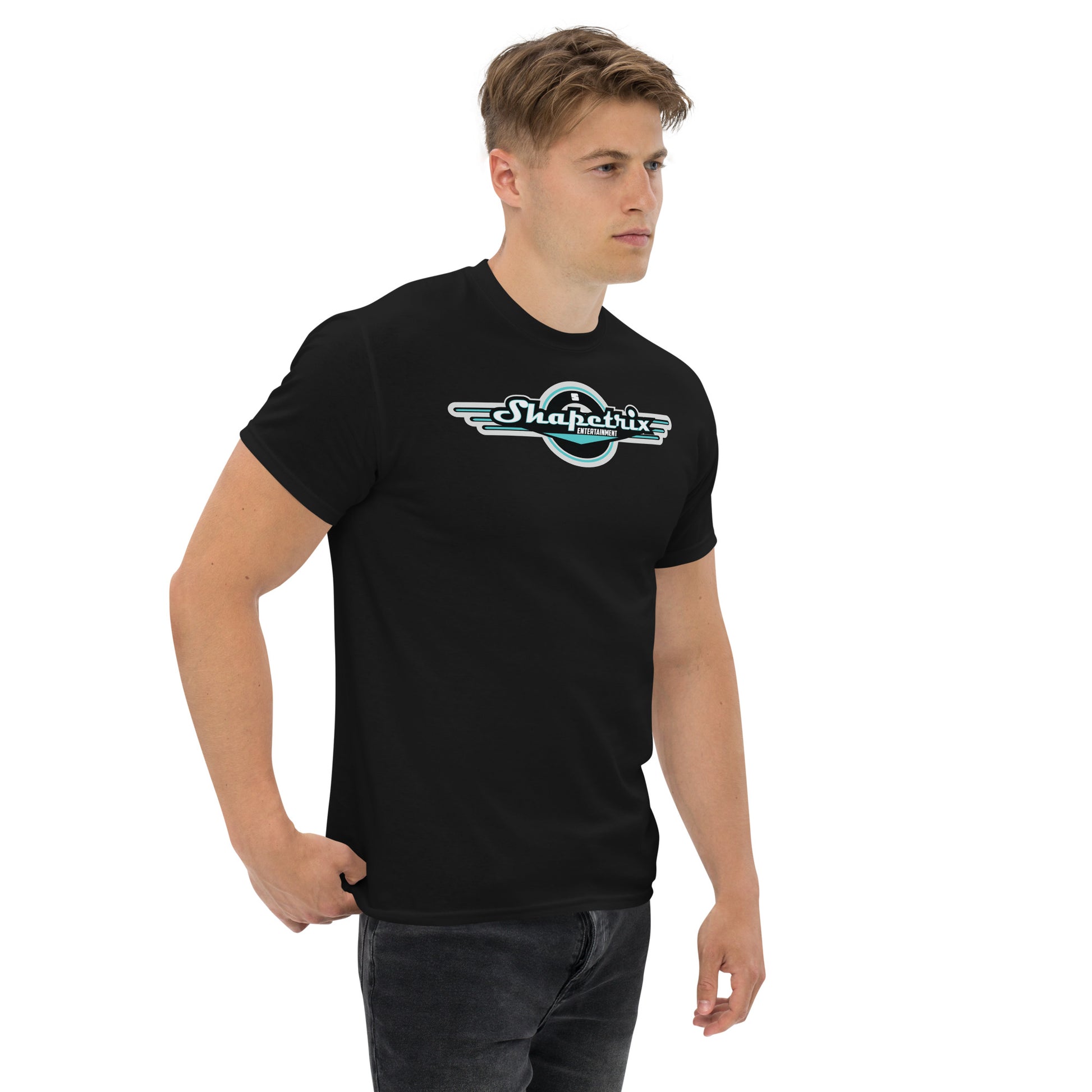 A young Caucasian man models a sleek black t-shirt featuring the Shapetrix Entertainment logo centered on the chest. The logo is designed with stylized teal and white text on a black circular emblem, suggesting a sense of motion with its wing-like extensions. The man stands casually with his body slightly turned to the side, giving a calm and composed look.