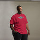 A smiling African American man wears a vibrant red t-shirt with the Shapetrix Entertainment logo in teal and white on the chest. The logo's dynamic, wing-like design on a black circular background adds a retro yet modern feel. The man's cheerful expression and relaxed pose, with one hand casually tucked into his jeans, create a friendly and approachable vibe.