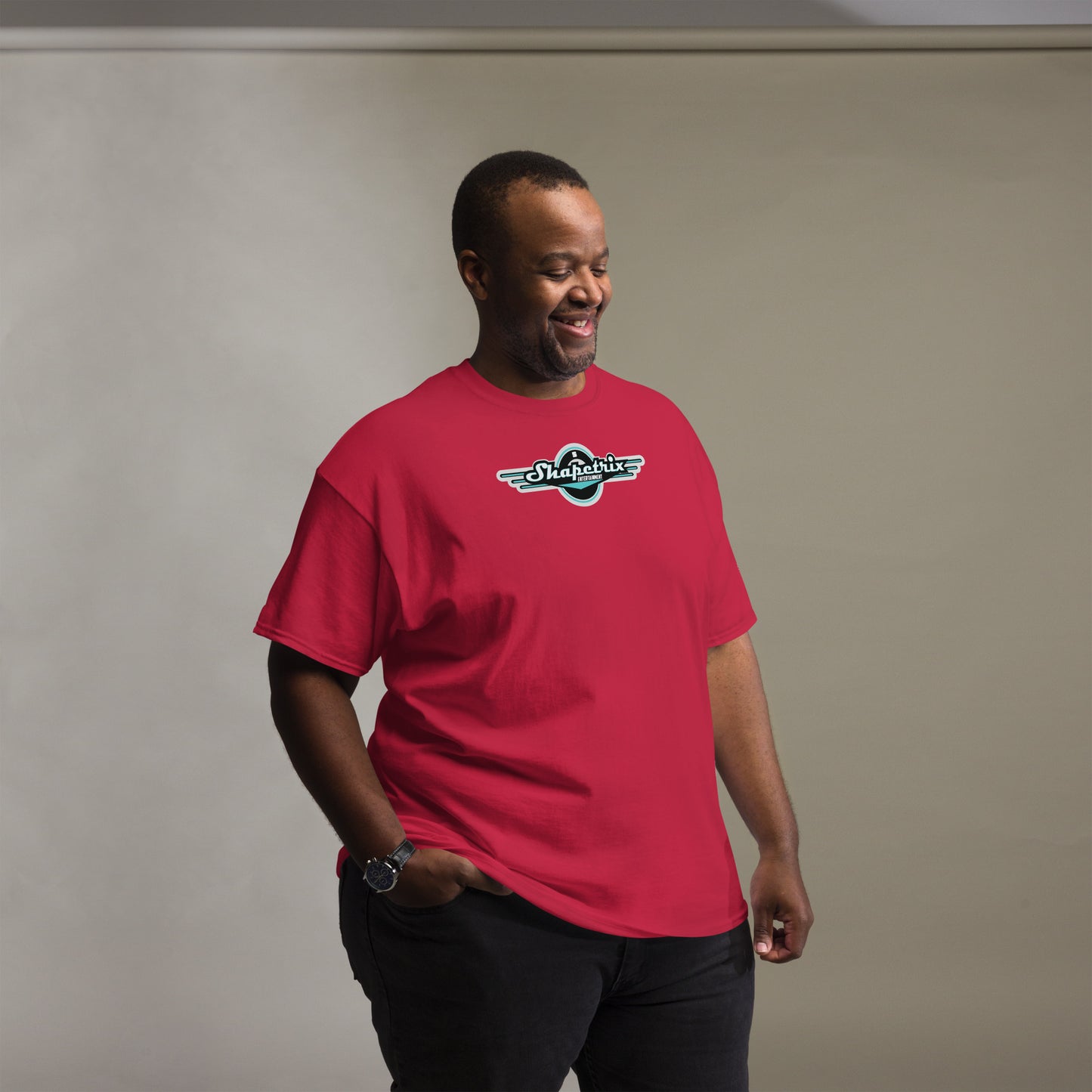 A smiling African American man wears a vibrant red t-shirt with the Shapetrix Entertainment logo in teal and white on the chest. The logo's dynamic, wing-like design on a black circular background adds a retro yet modern feel. The man's cheerful expression and relaxed pose, with one hand casually tucked into his jeans, create a friendly and approachable vibe.