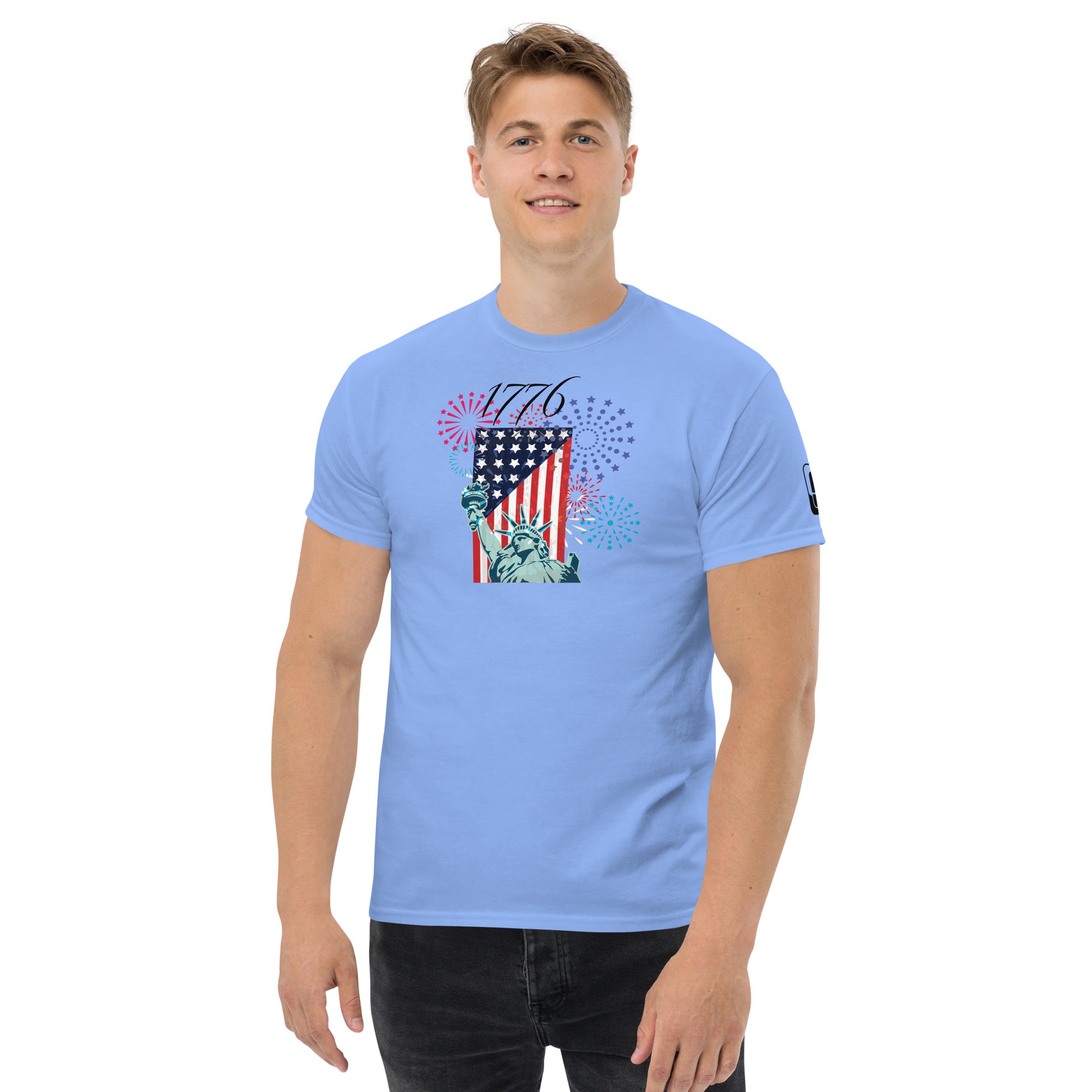 A young man wearing a Carolina blue t-shirt with a festive Fourth of July design. The graphic shows the Statue of Liberty and an American flag, overlaid with colorful fireworks in red, blue, and purple bursts. The design celebrates American independence with '1776' boldly emblazoned across the top.
