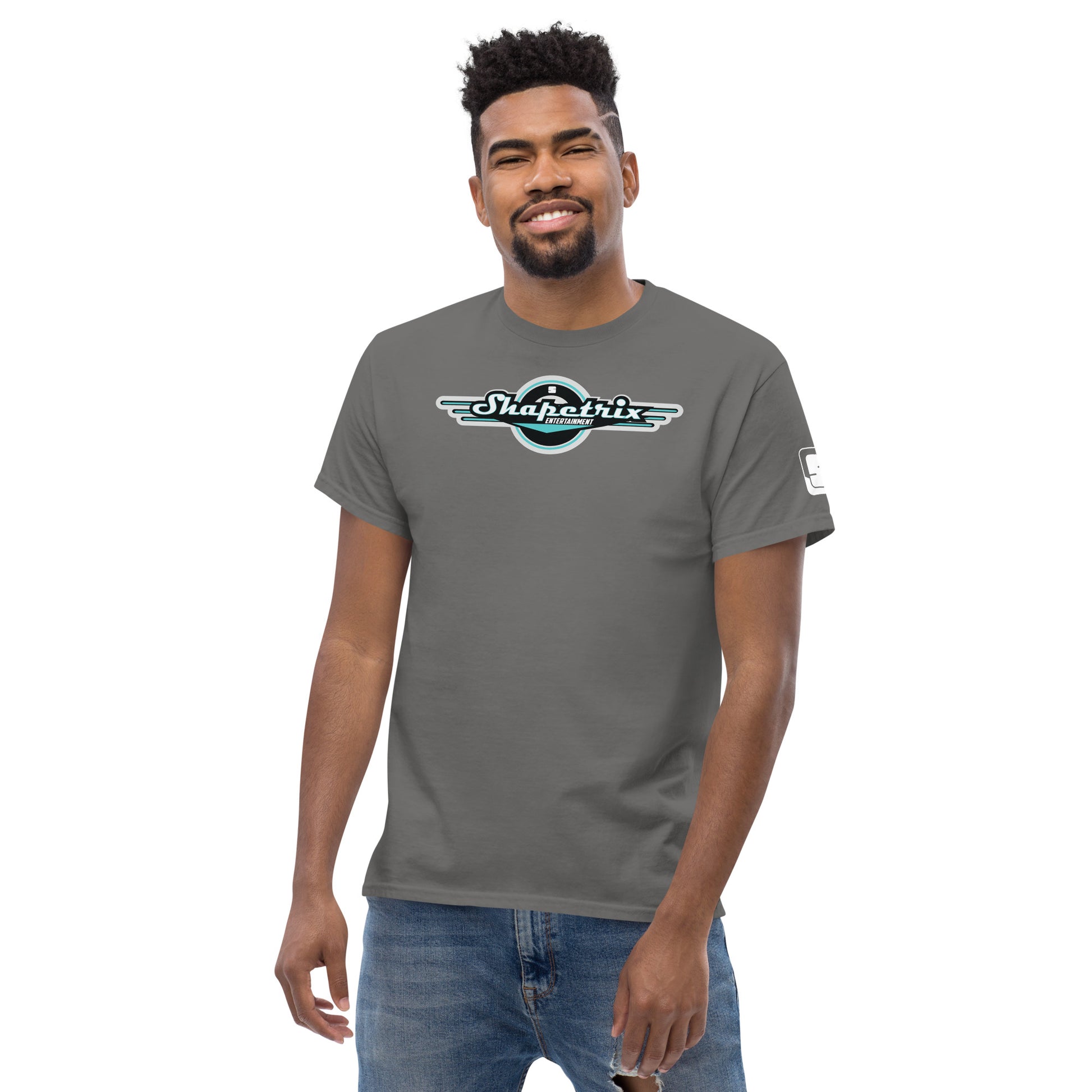 A cheerful African-American man standing confidently with a subtle smile, wearing a Shapetrix Entertainment t-shirt in a cool gray shade. The shirt features a prominently displayed logo with teal and white lettering accentuated by a stylized wing design on a black circular background. His casual look is completed with light blue jeans, and he exudes an upbeat and friendly vibe.