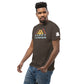 A young African American man poses casually, looking slightly to the side with a soft, confident smile. He wears a dark brown t-shirt emblazoned with the "Happy Camper" design, which features an abstract mountain design in muted colors of yellow and orange above the phrase in pale blue, sans-serif font. The outfit is paired with distressed blue jeans, contributing to his stylish and relaxed demeanor.