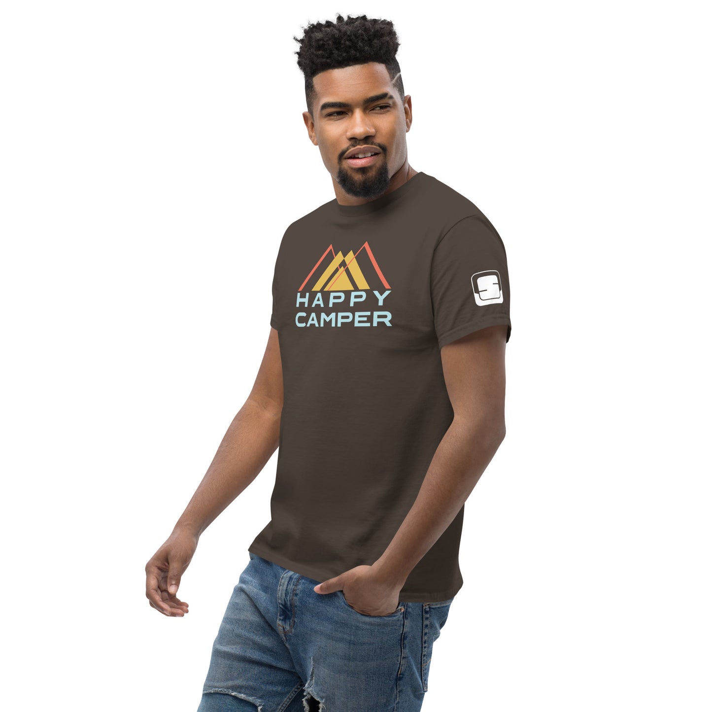 A young African American man poses casually, looking slightly to the side with a soft, confident smile. He wears a dark brown t-shirt emblazoned with the "Happy Camper" design, which features an abstract mountain design in muted colors of yellow and orange above the phrase in pale blue, sans-serif font. The outfit is paired with distressed blue jeans, contributing to his stylish and relaxed demeanor.